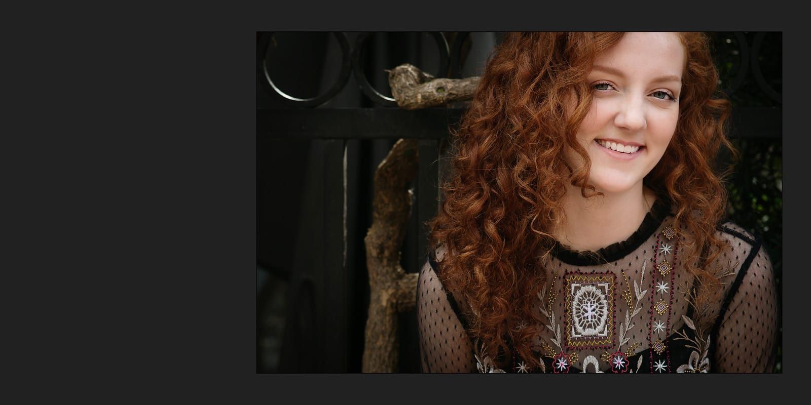 A smiling woman with curly red hair, wearing a sheer top, posing outside.
