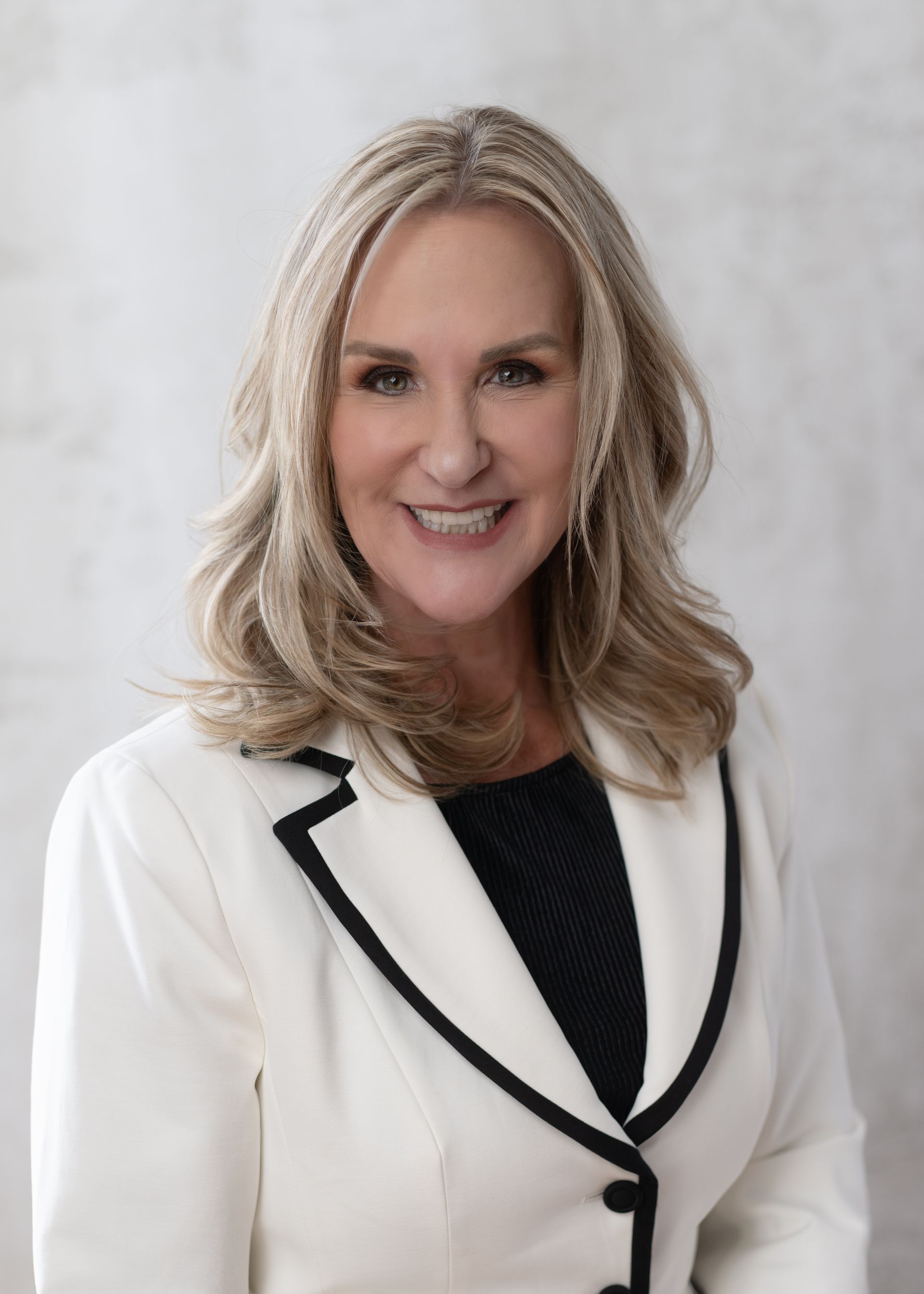 Woman in a white blazer with black trim smiles, her blonde hair shoulder-length, against a neutral background.