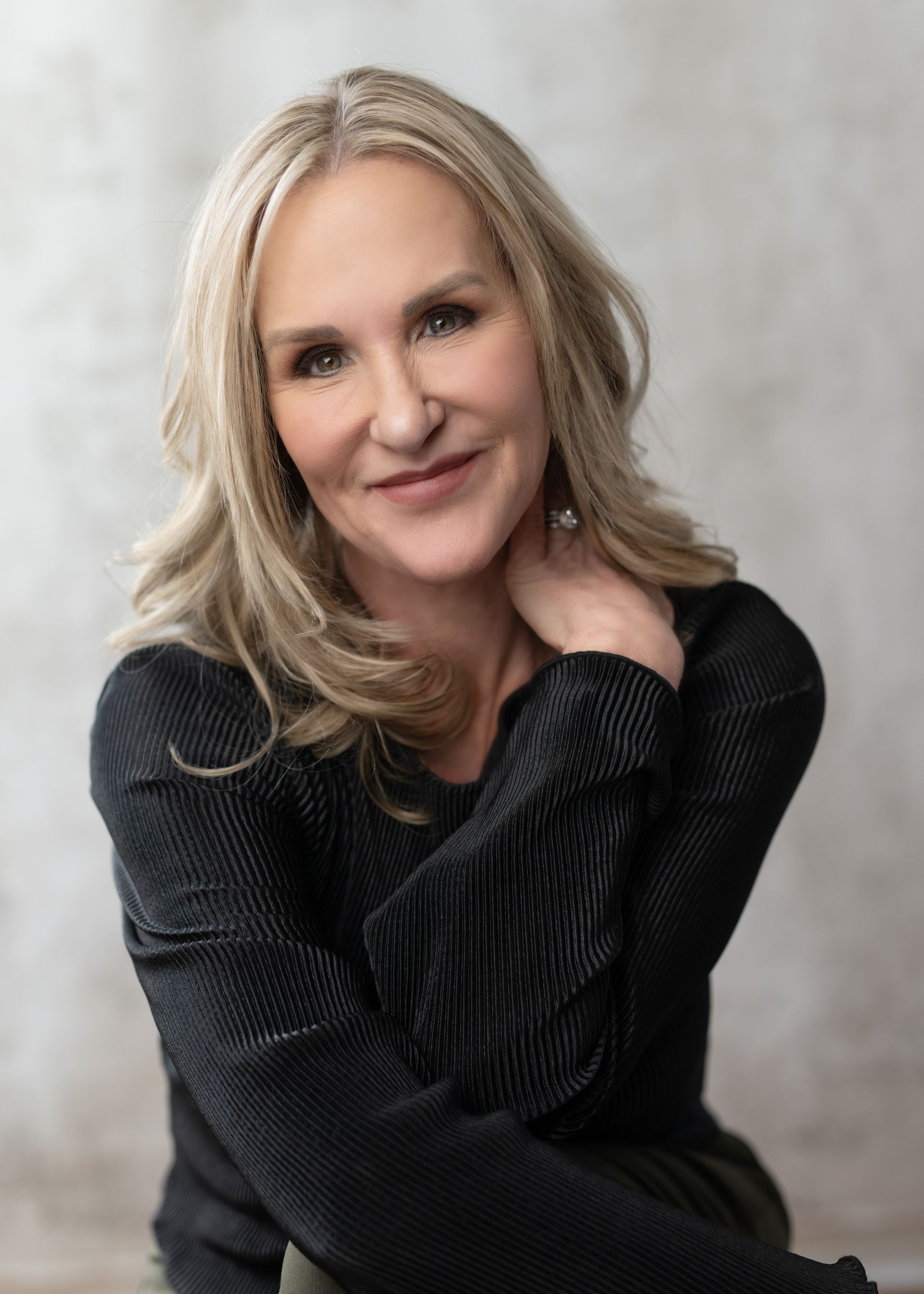 Blonde woman with a gentle smile, black sweater, and hand on neck, seated against a neutral backdrop.