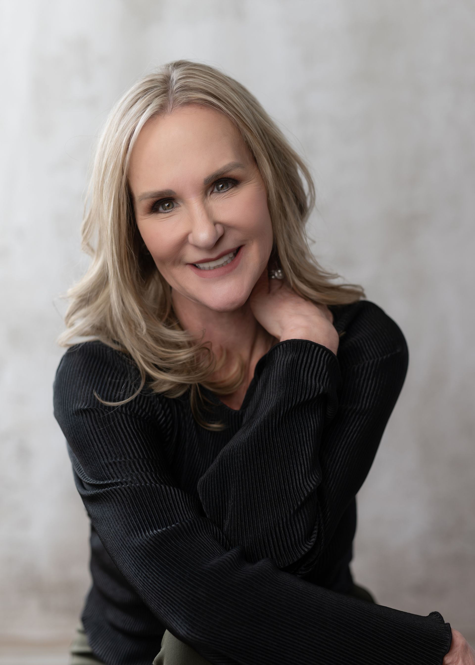 Blonde woman with hand on neck smiles at the camera, wearing a black sweater, against a light gray backdrop.