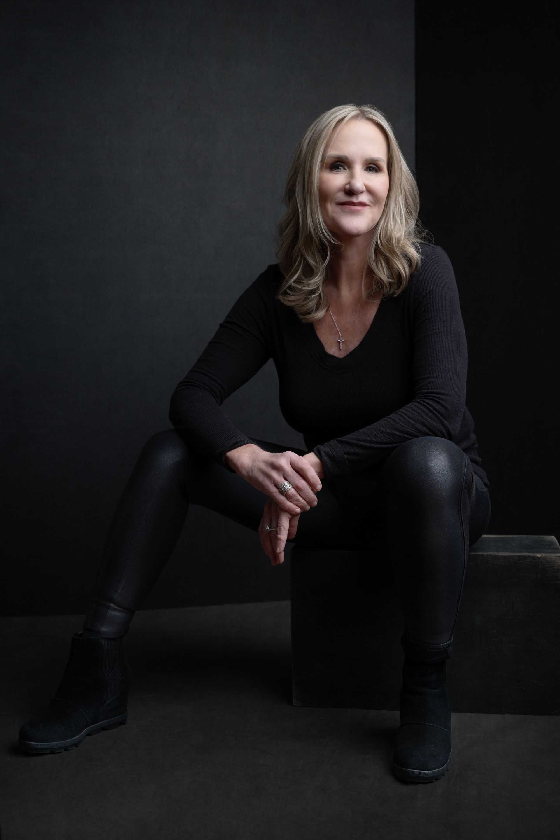 Woman with blonde hair, smiles sitting on a dark block, wearing black top, leather pants, and boots.