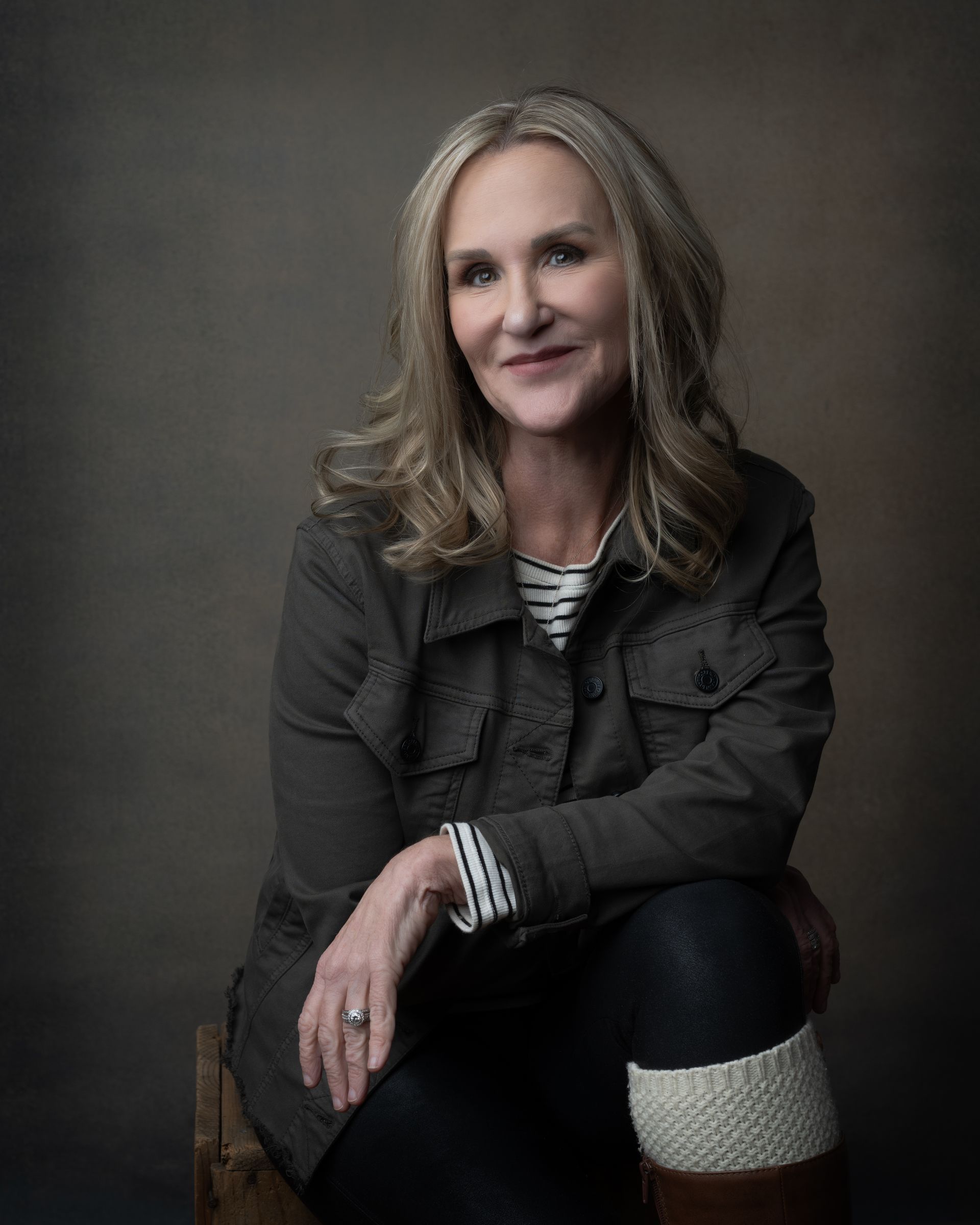 Woman with graying hair, wearing a jacket and boots, smiles at the camera. Neutral background.