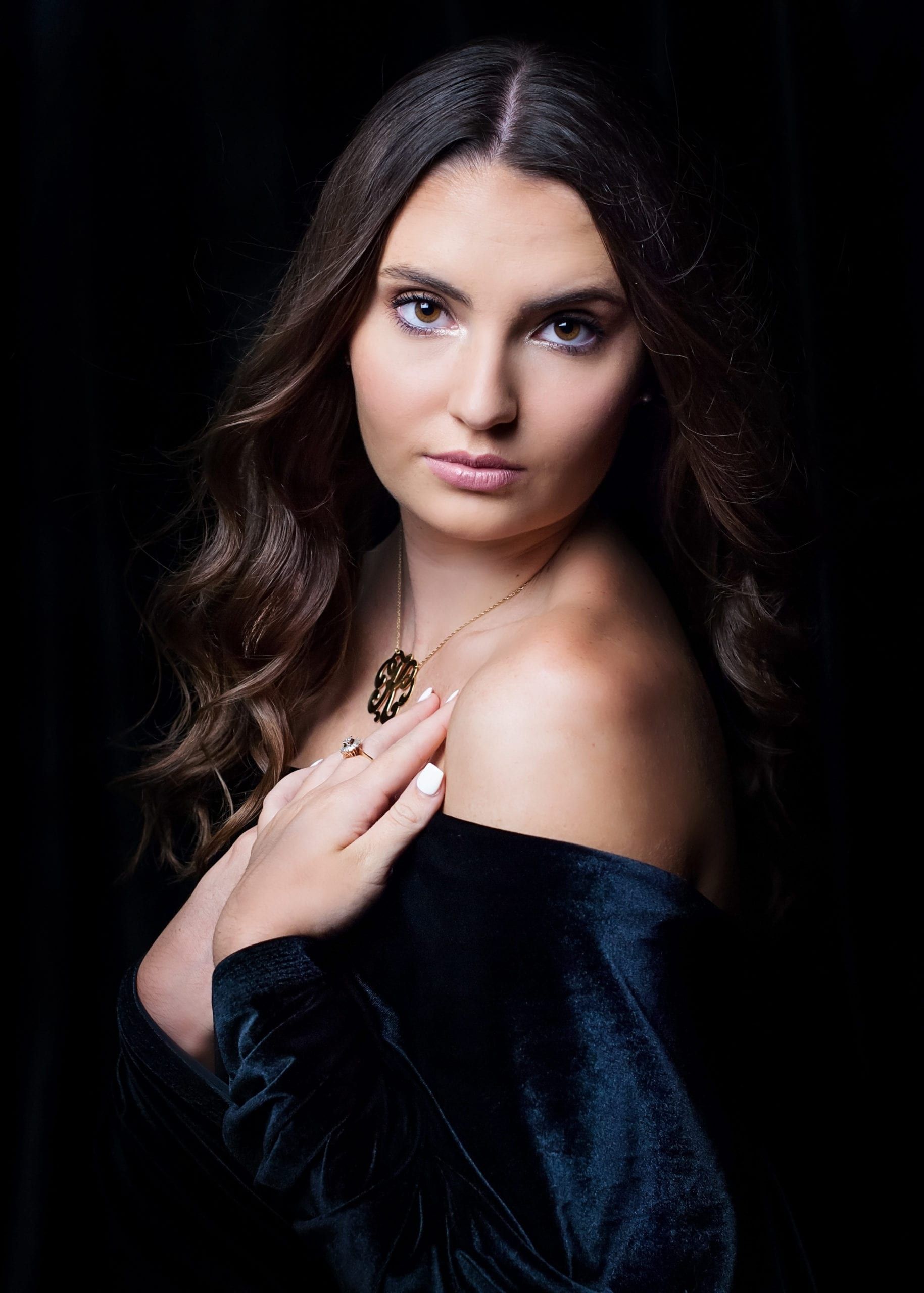 Woman with long wavy brown hair, wearing an off-shoulder dark velvet top, looking at the viewer.