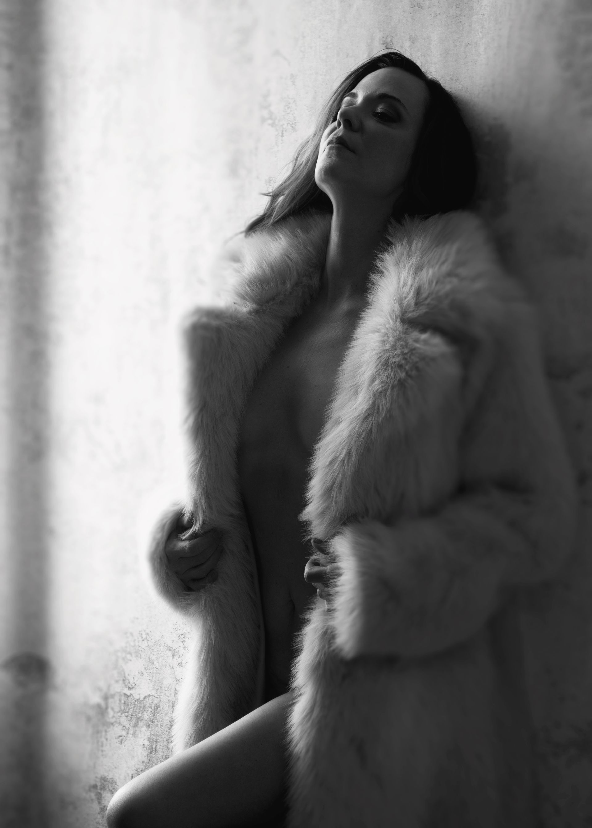 Woman in a fur coat, leaning against a wall with eyes closed. Black and white.