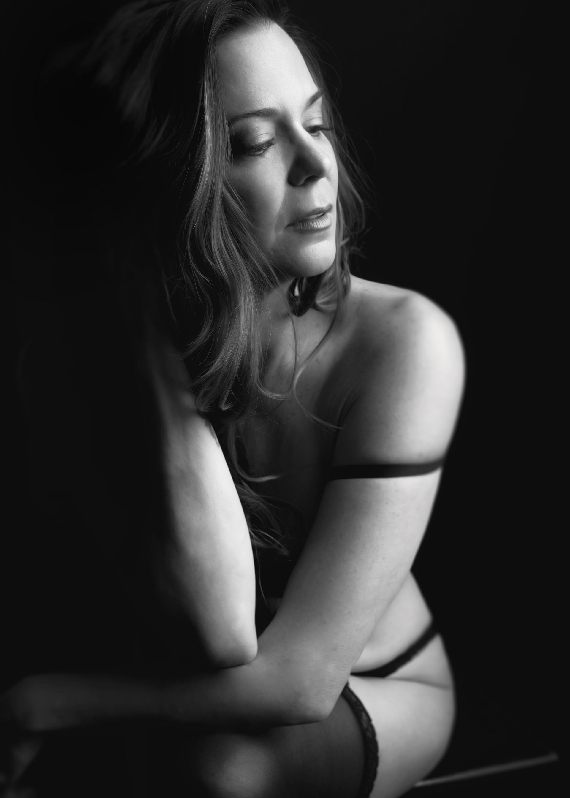 Black and white photo of a woman in lingerie, eyes closed, arms crossed, dark background.
