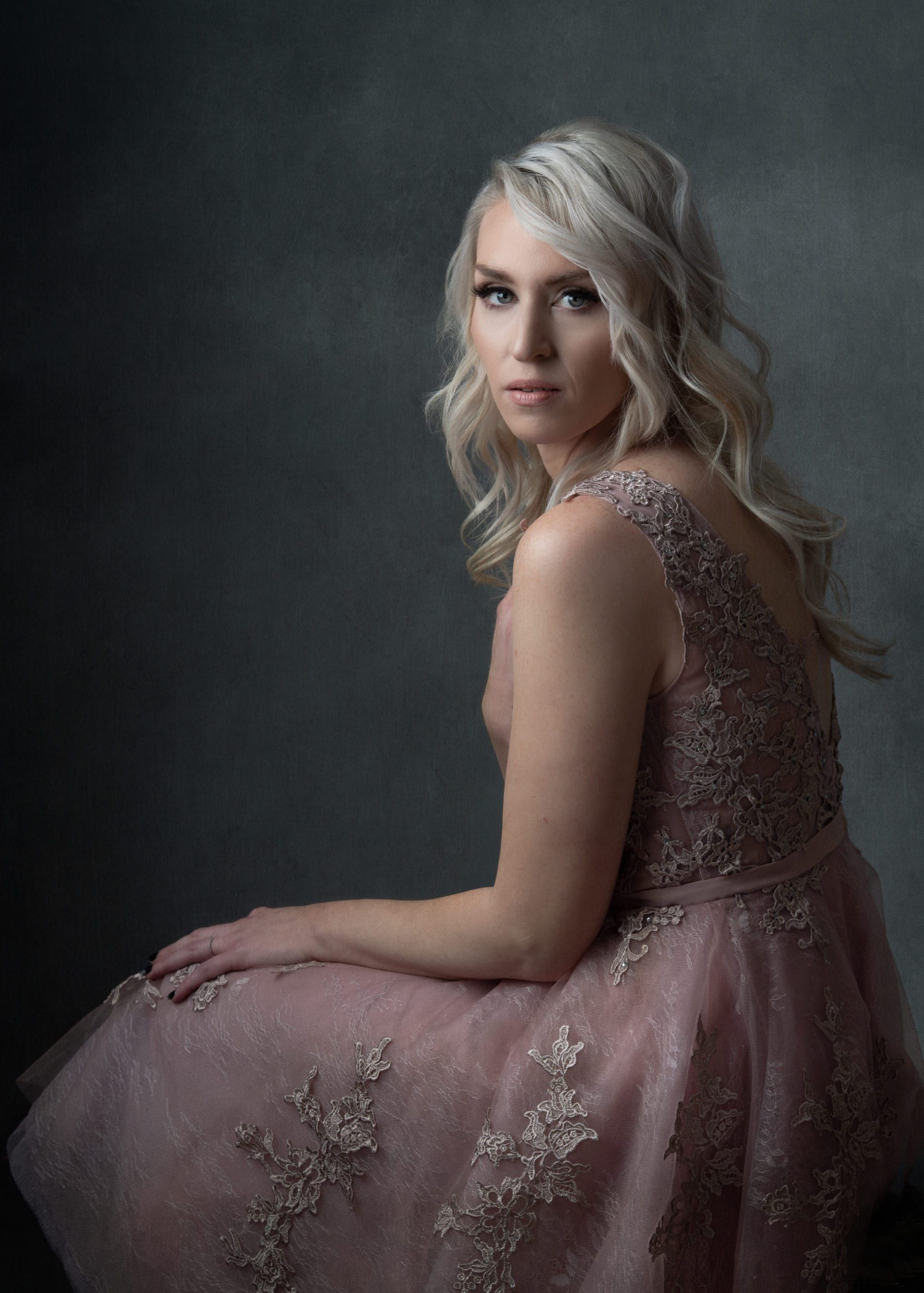 Blonde woman in pink gown poses, looking towards the viewer. Dark grey backdrop.
