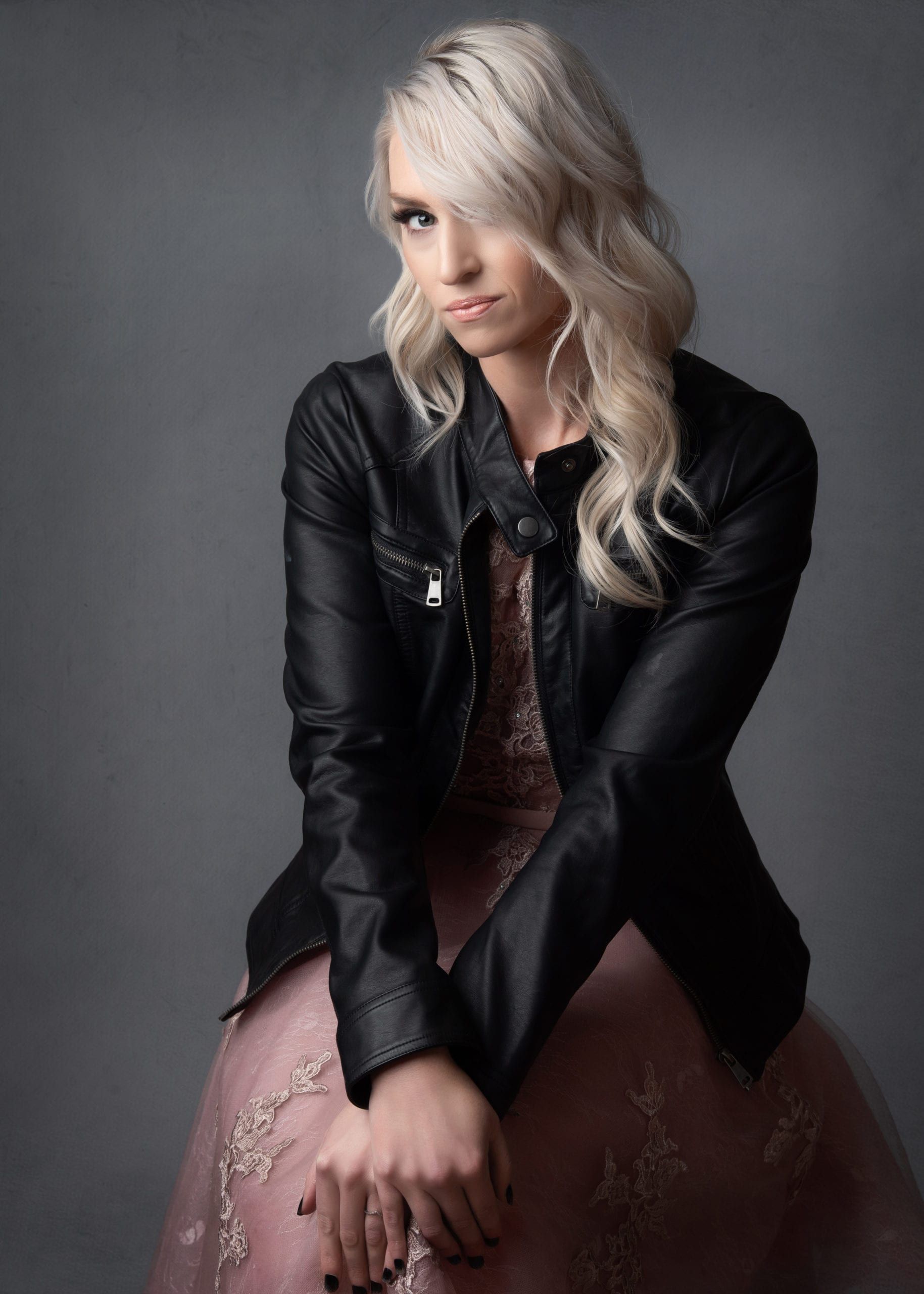 Woman in a pink dress and black jacket, blonde hair, looking at the camera. Gray background.