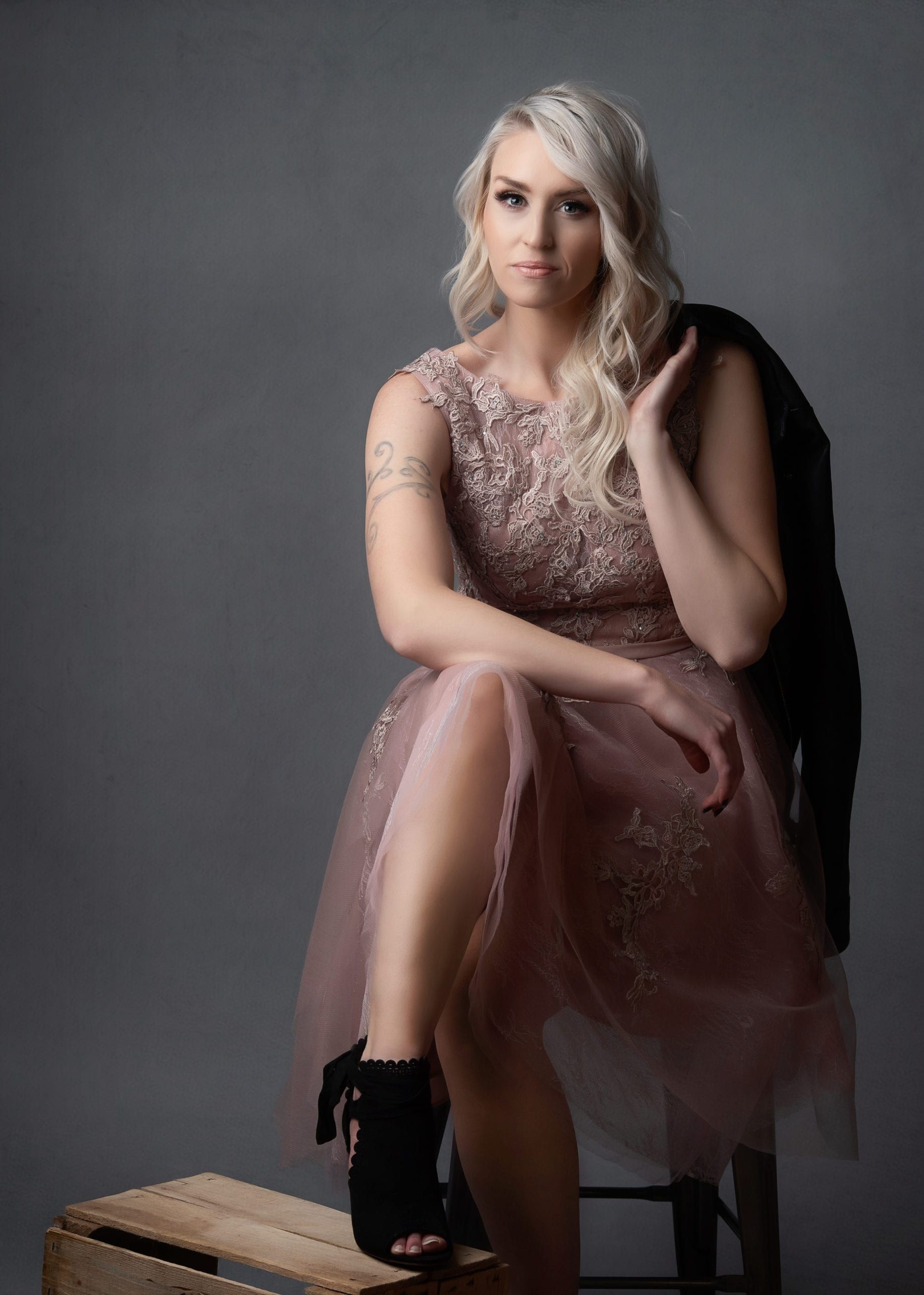 Blonde woman in pink dress poses, seated, with jacket; gray background.