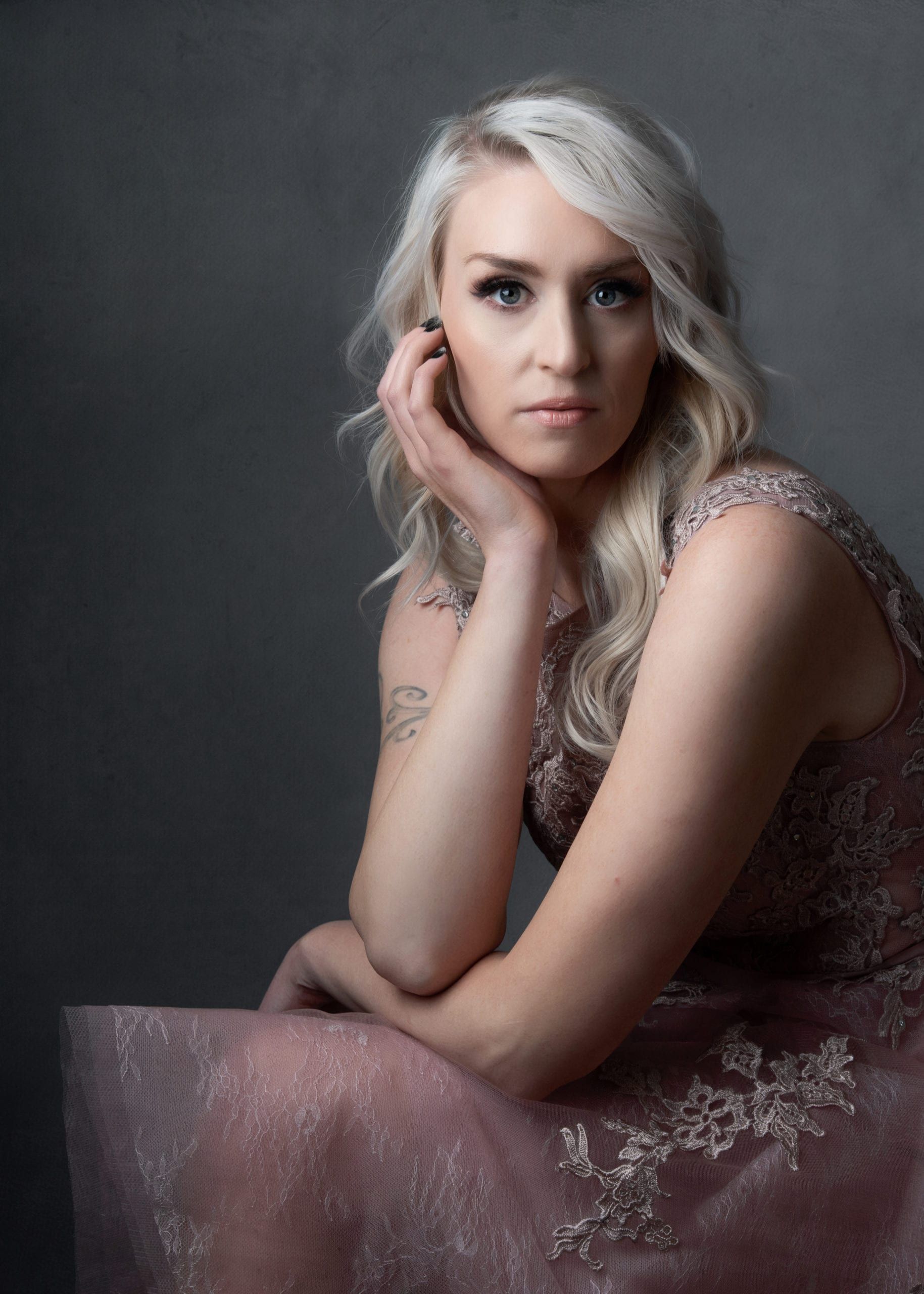 Blonde woman in pink dress rests chin on hand, looking at the camera against a gray backdrop.