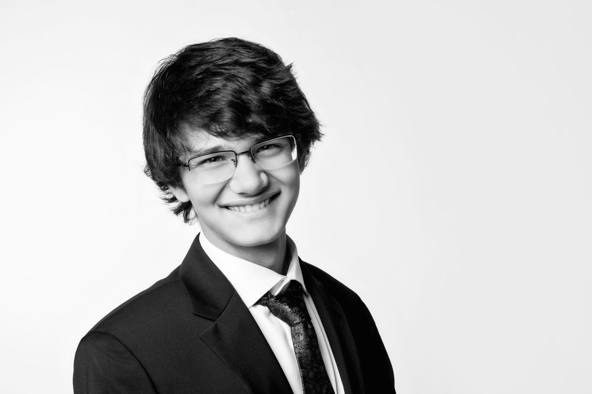 Smiling young man with glasses, dark hair, and suit and tie, against a white background.