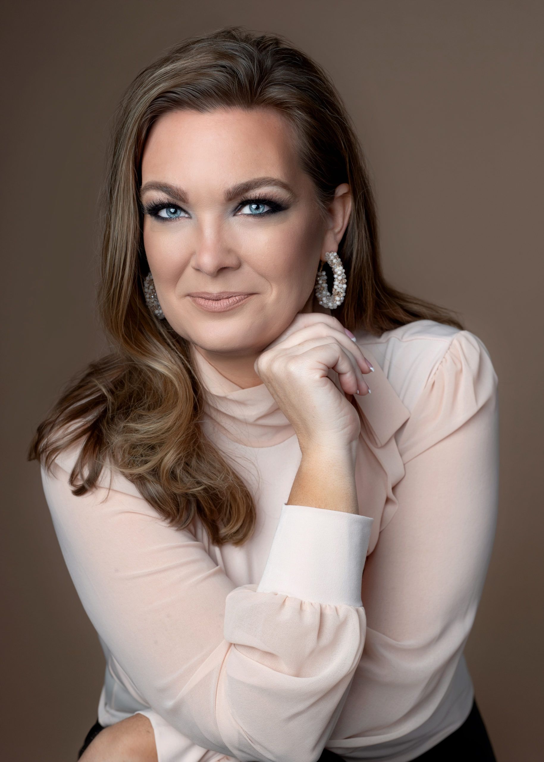 Woman with long brown hair, blue eyes, wearing a blush pink top and large hoop earrings, smiles, posing in front of a light brown backdrop.