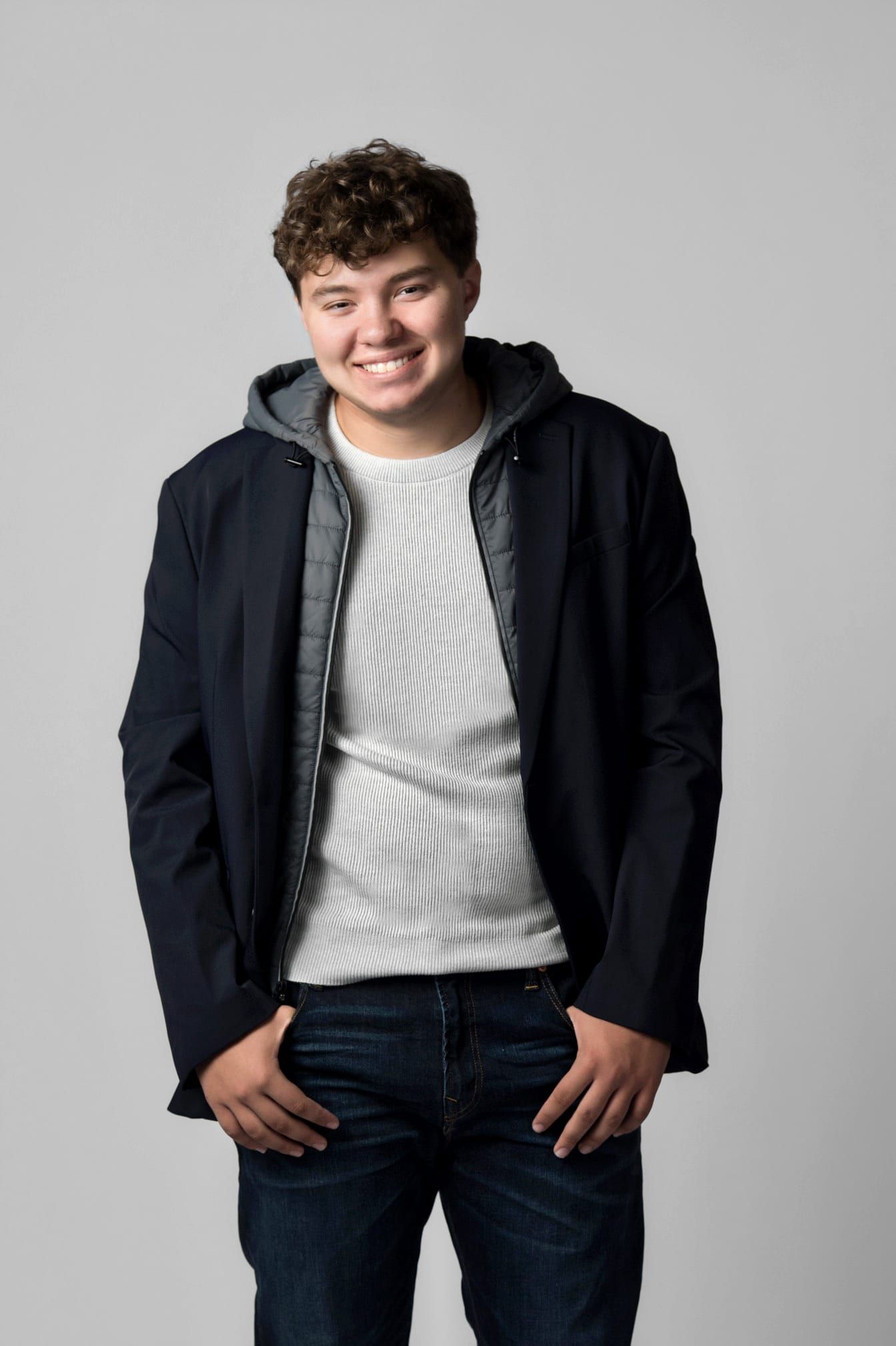 Young man with curly hair smiles, wearing a striped shirt, blue jacket, dark jeans, hands in pockets.