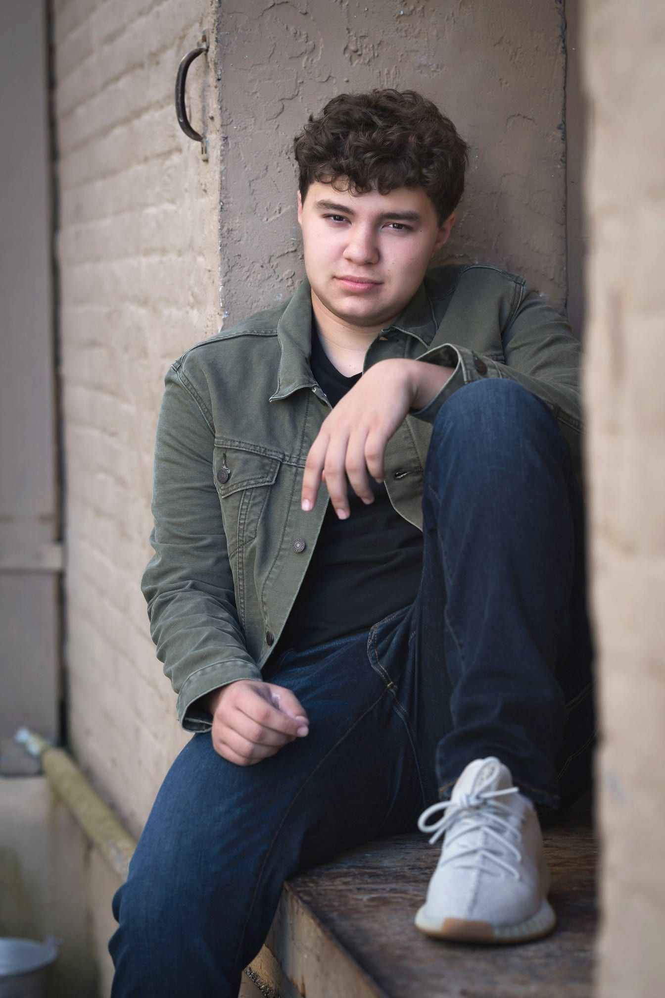 Young man sits outside wearing jeans and a green jacket.