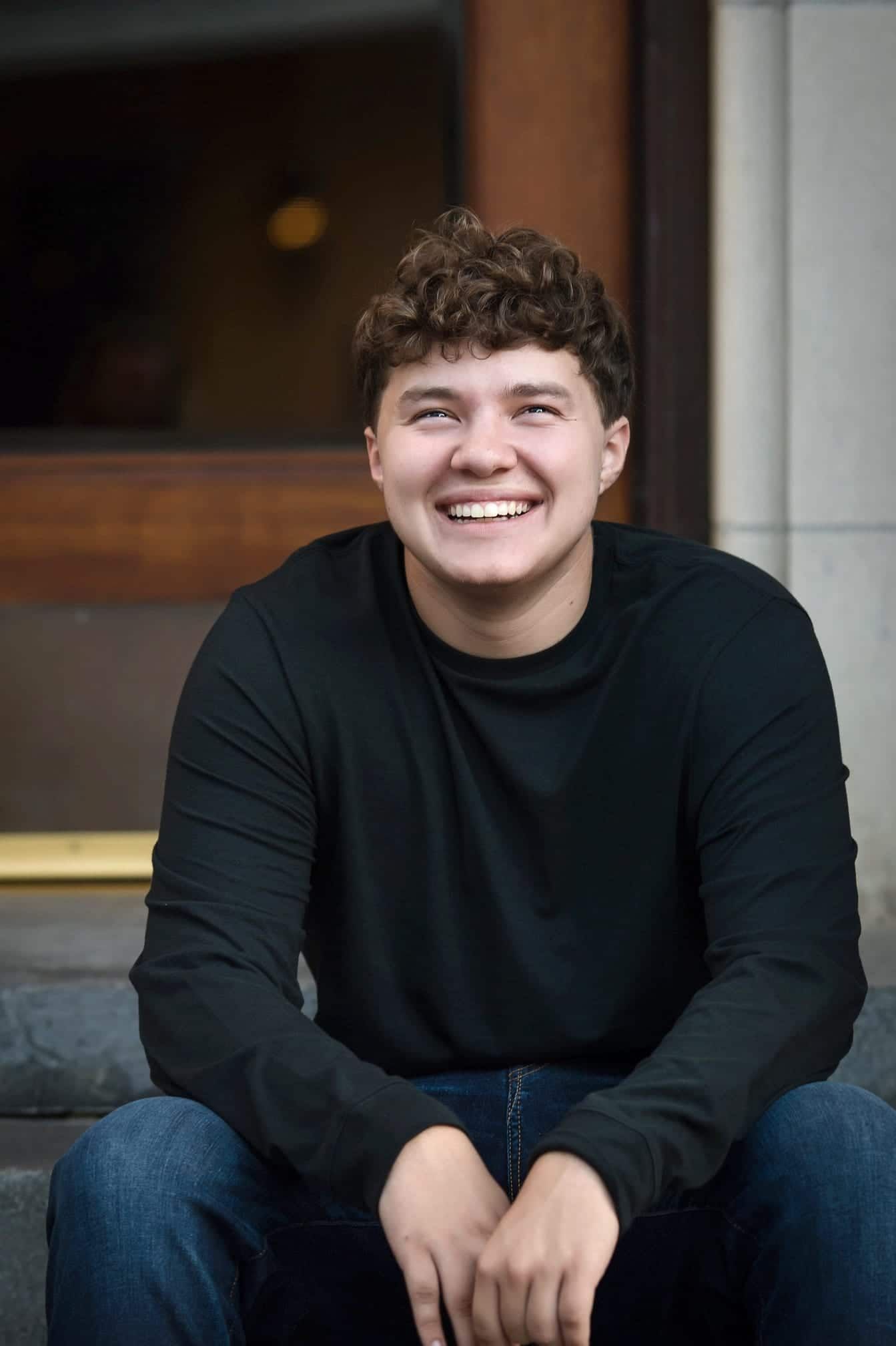 Young man with curly hair smiles broadly while sitting on steps; wearing black long-sleeve shirt and jeans.