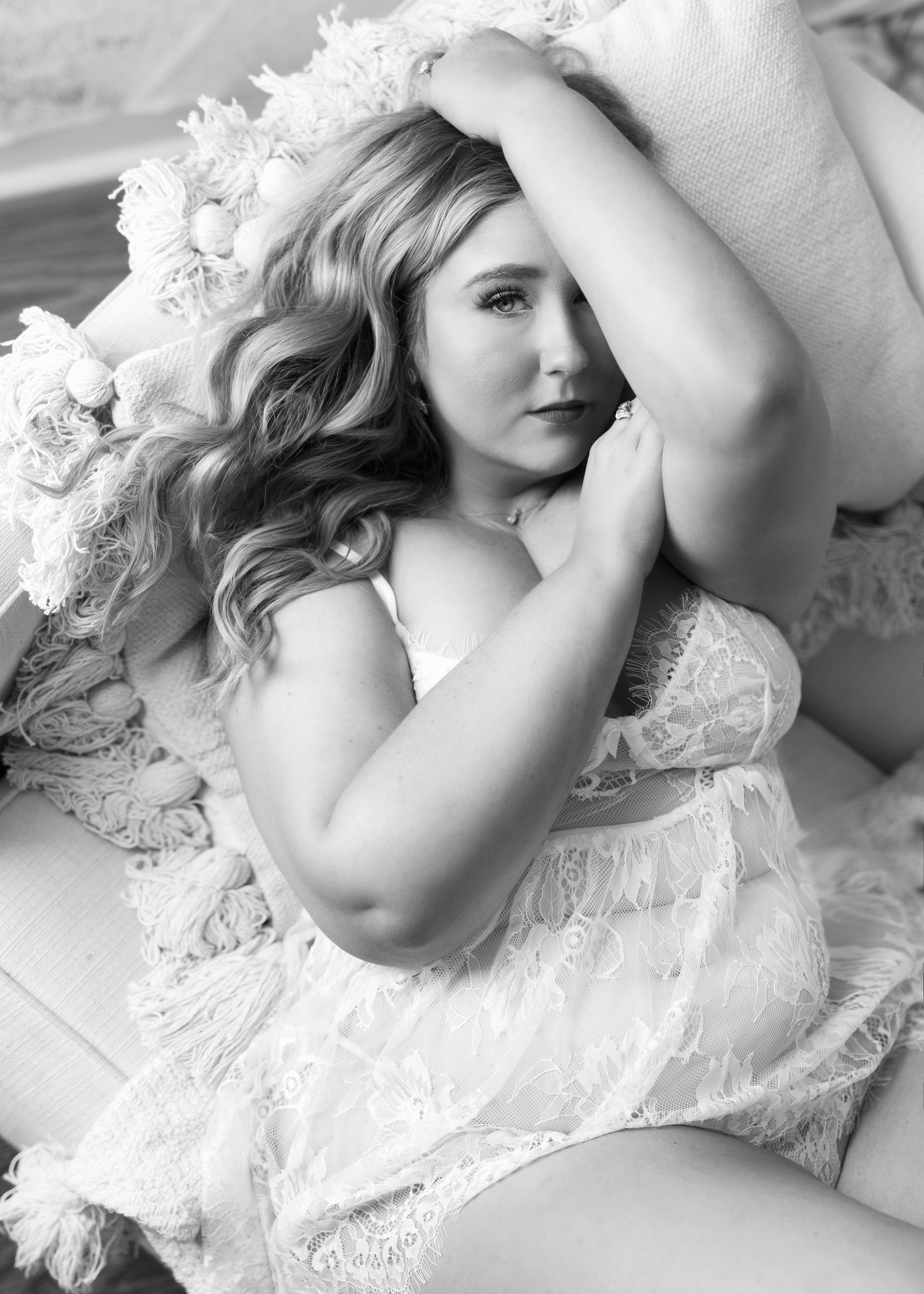 Woman in lace lingerie reclines, looking at the camera, in a soft, neutral-toned setting.
