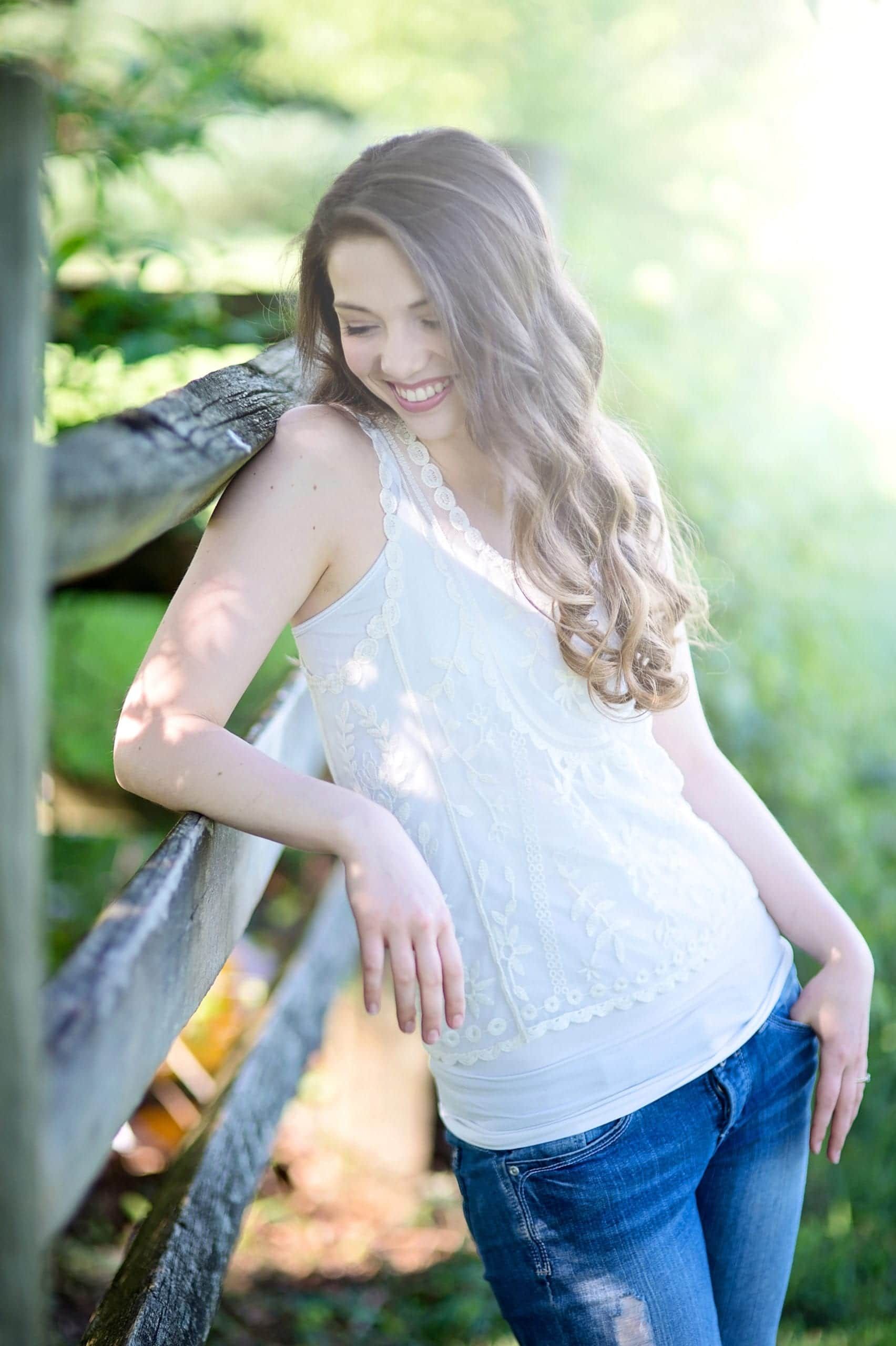 Woman in white top and jeans leaning against a wooden fence, smiling outdoors.