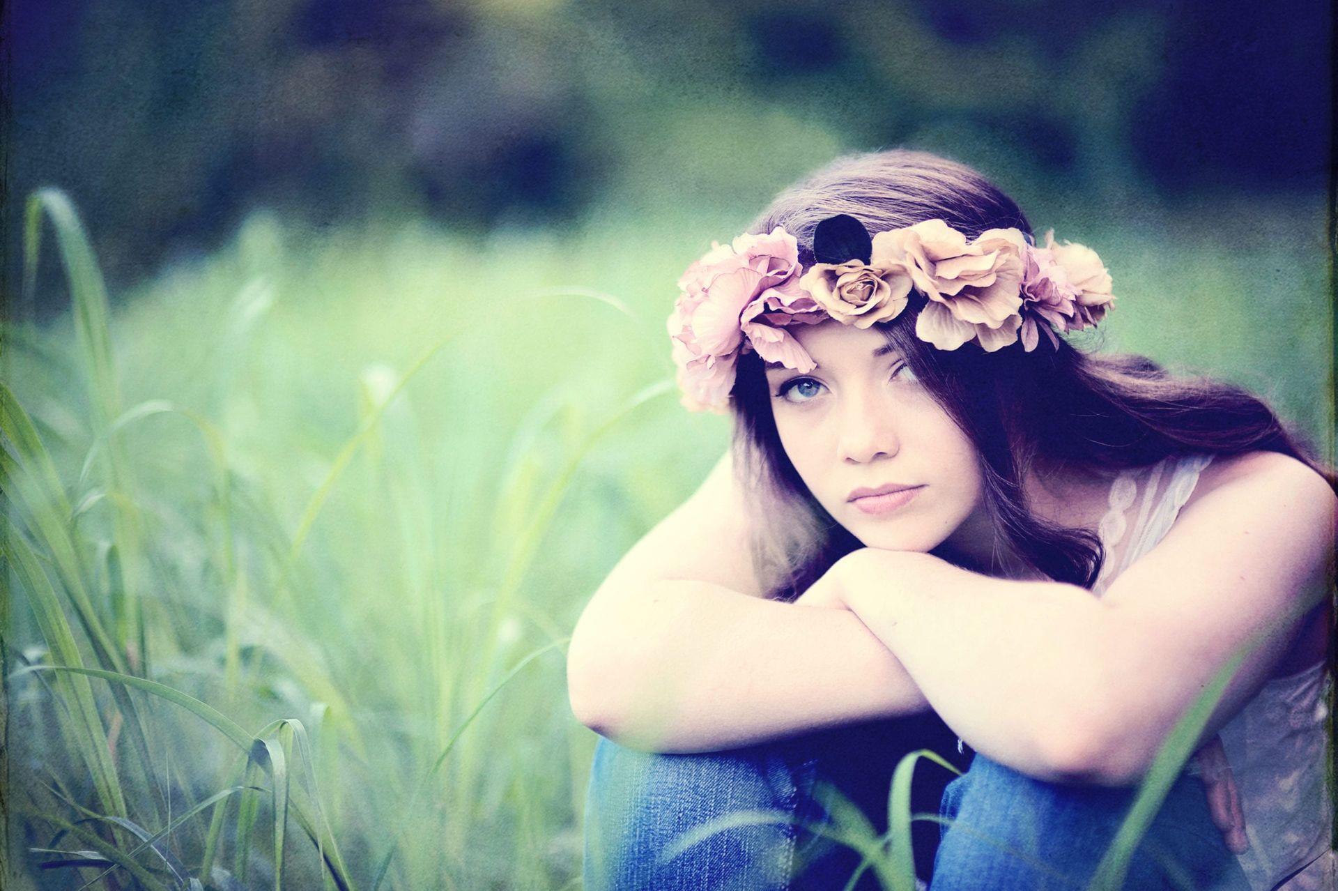 Woman wearing a flower crown, sitting in tall grass, looking pensive, with a soft, dreamy focus.