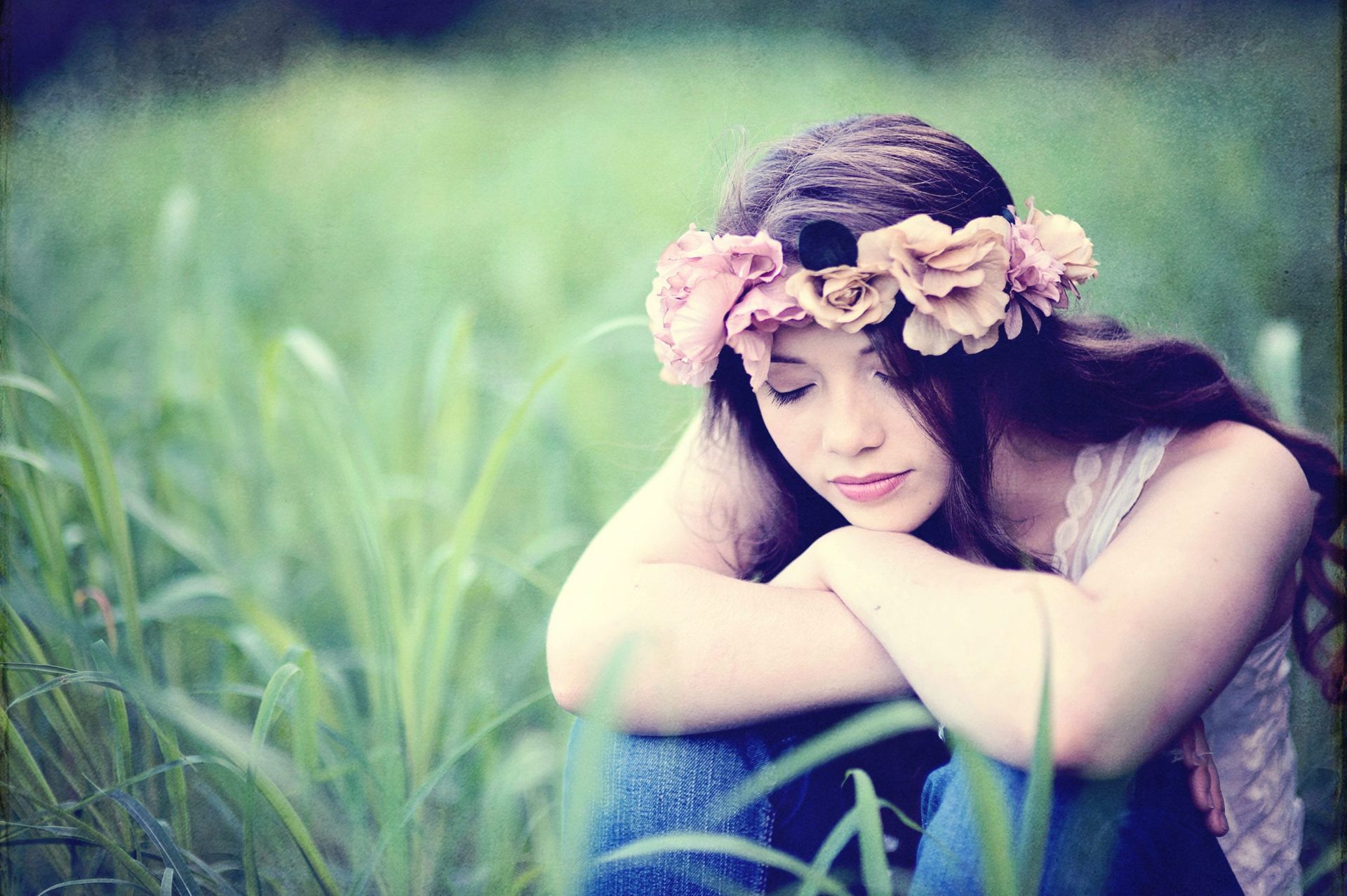 Woman with flower crown sitting in a field, looking down, arms crossed, surrounded by green grass.