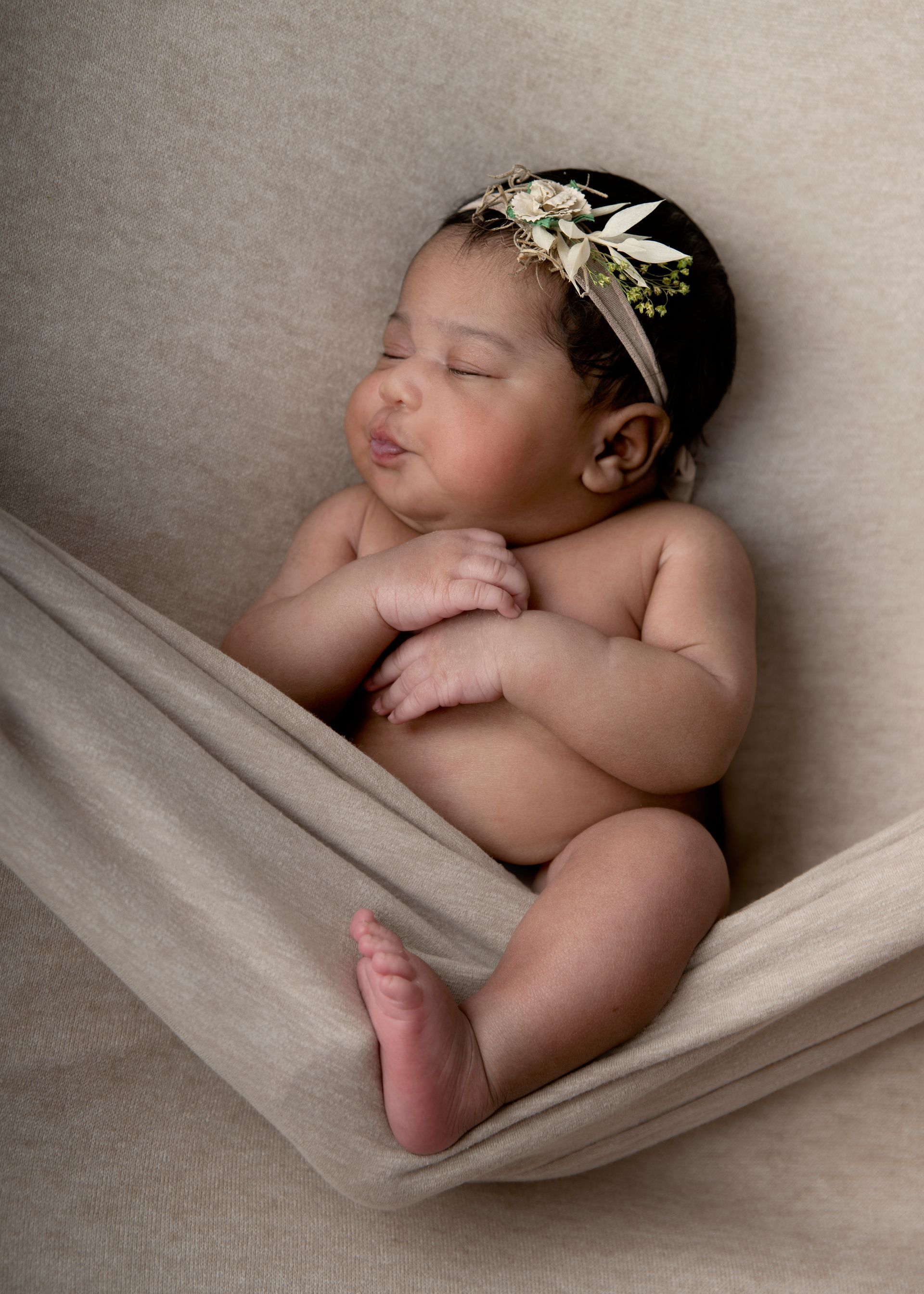 Newborn baby sleeping in a beige fabric hammock, wearing a floral headband.