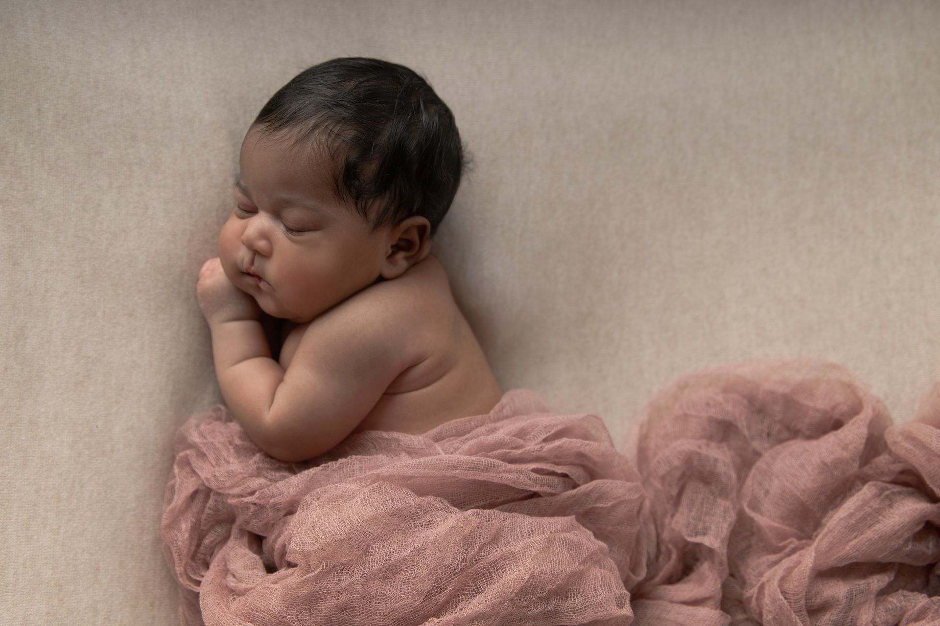 Newborn baby sleeping, wrapped in pink fabric, on a neutral beige surface.