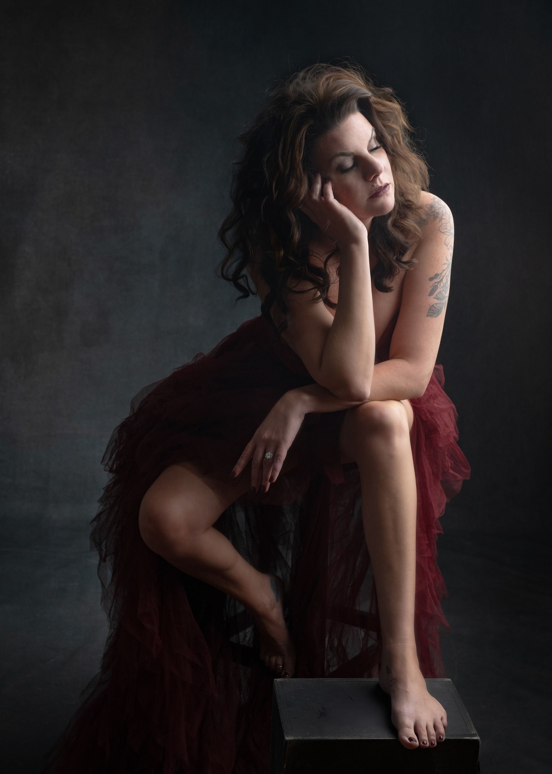 Woman draped in red fabric, leaning on a dark box, gazing down thoughtfully. Dark background.