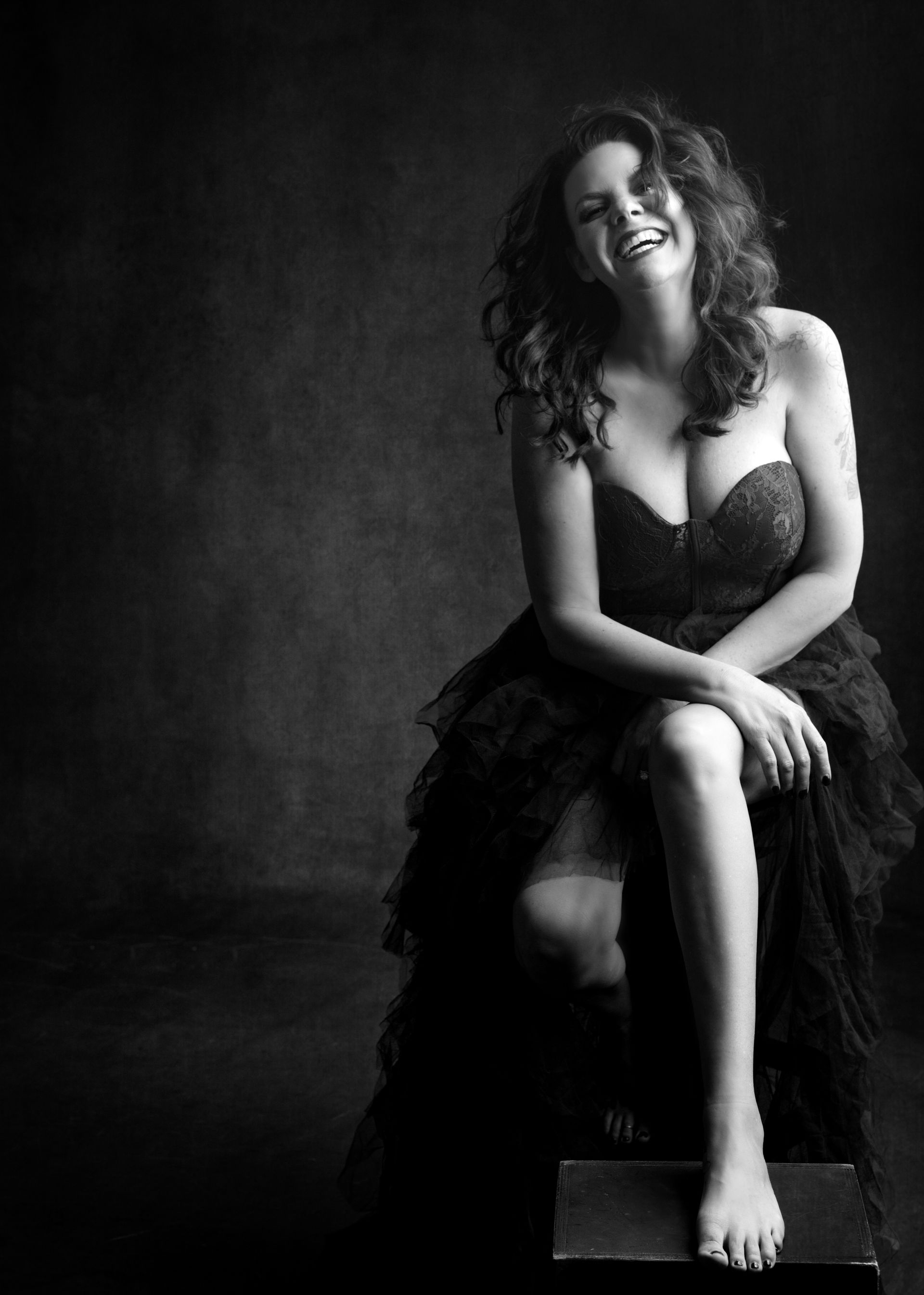 Woman in black gown, laughing, sitting on a box. Black and white portrait, dark background.