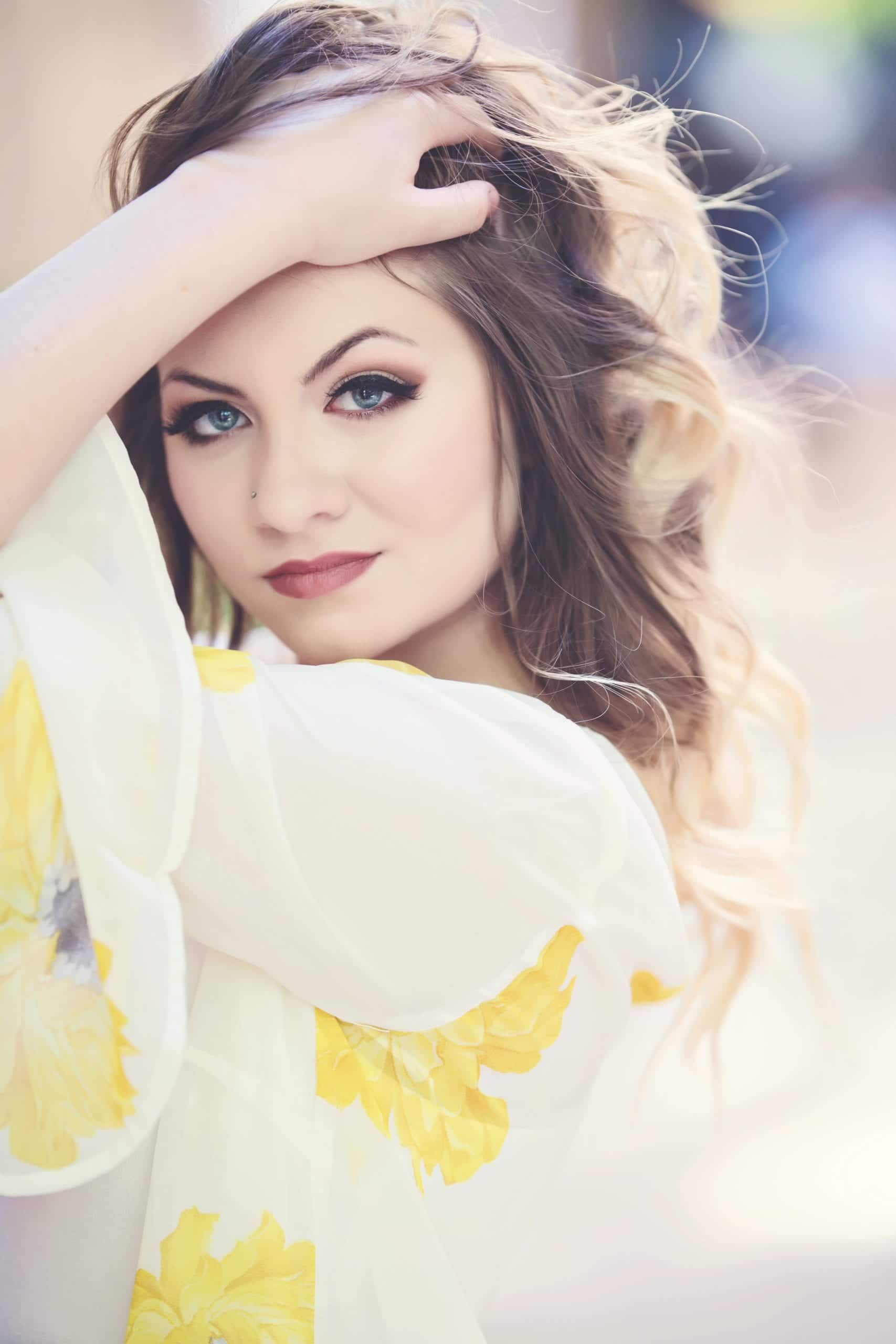 Woman with wavy, highlighted hair, blue eyes, and bold makeup, wearing a white blouse with yellow floral accents, hand in hair.