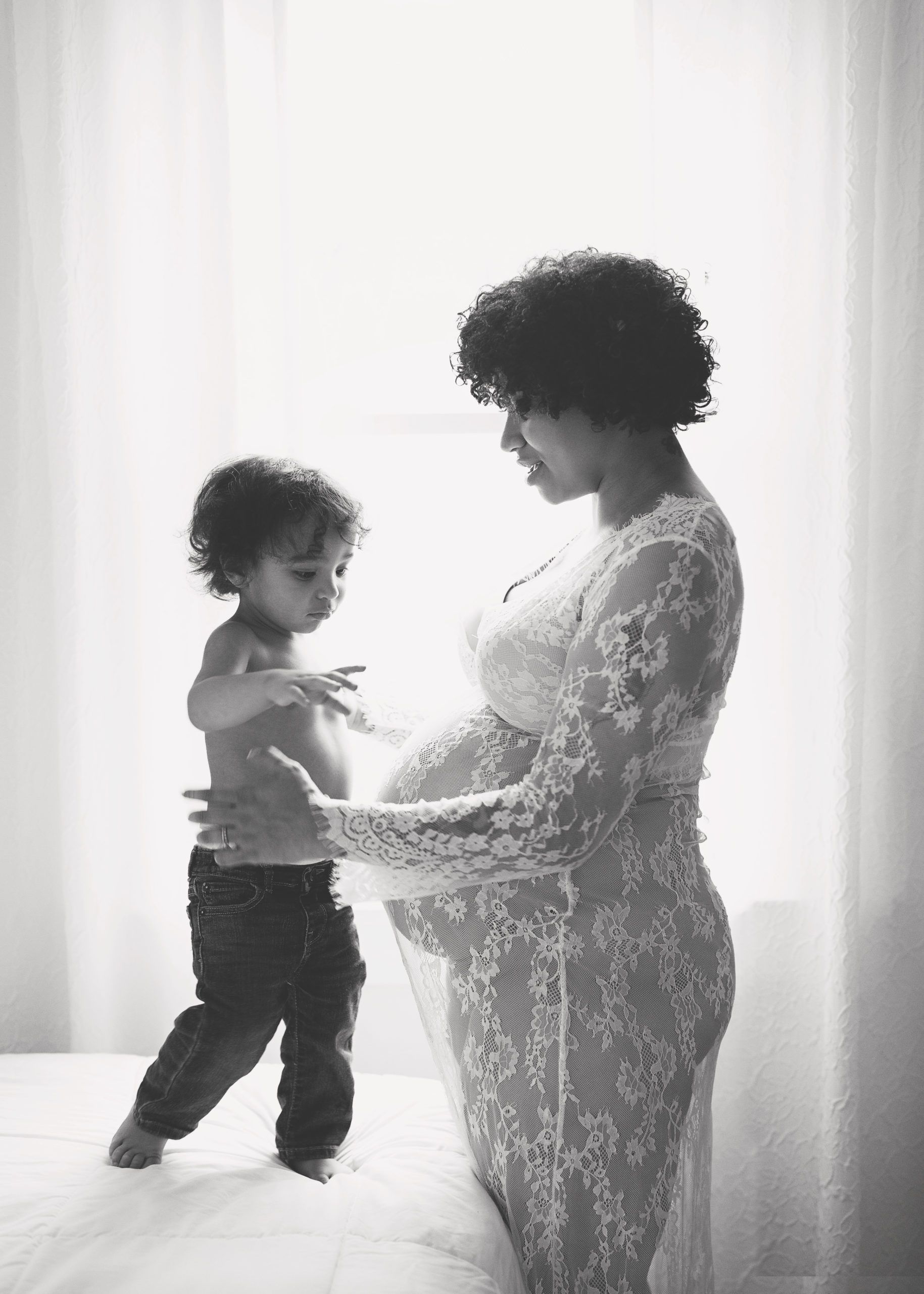 Pregnant woman in lace dress holds belly as toddler stands beside her. Black and white photo.