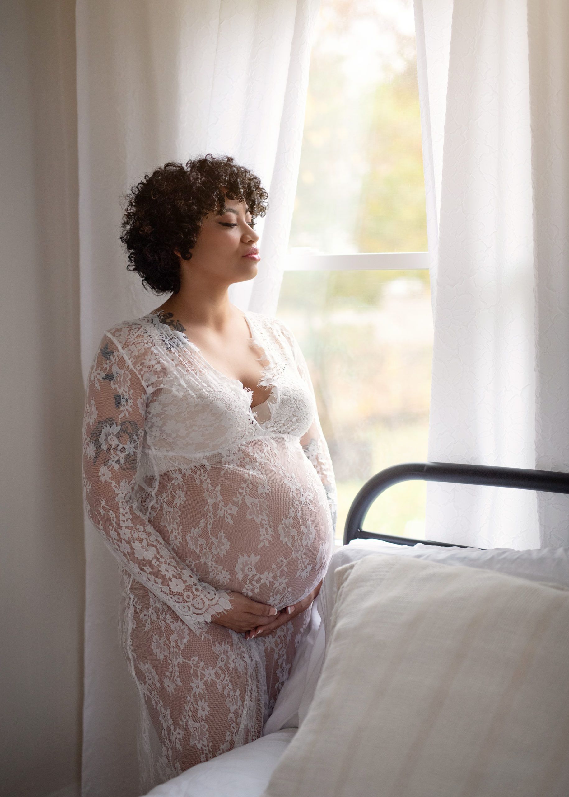 Pregnant woman in lace robe near a window, looking out.