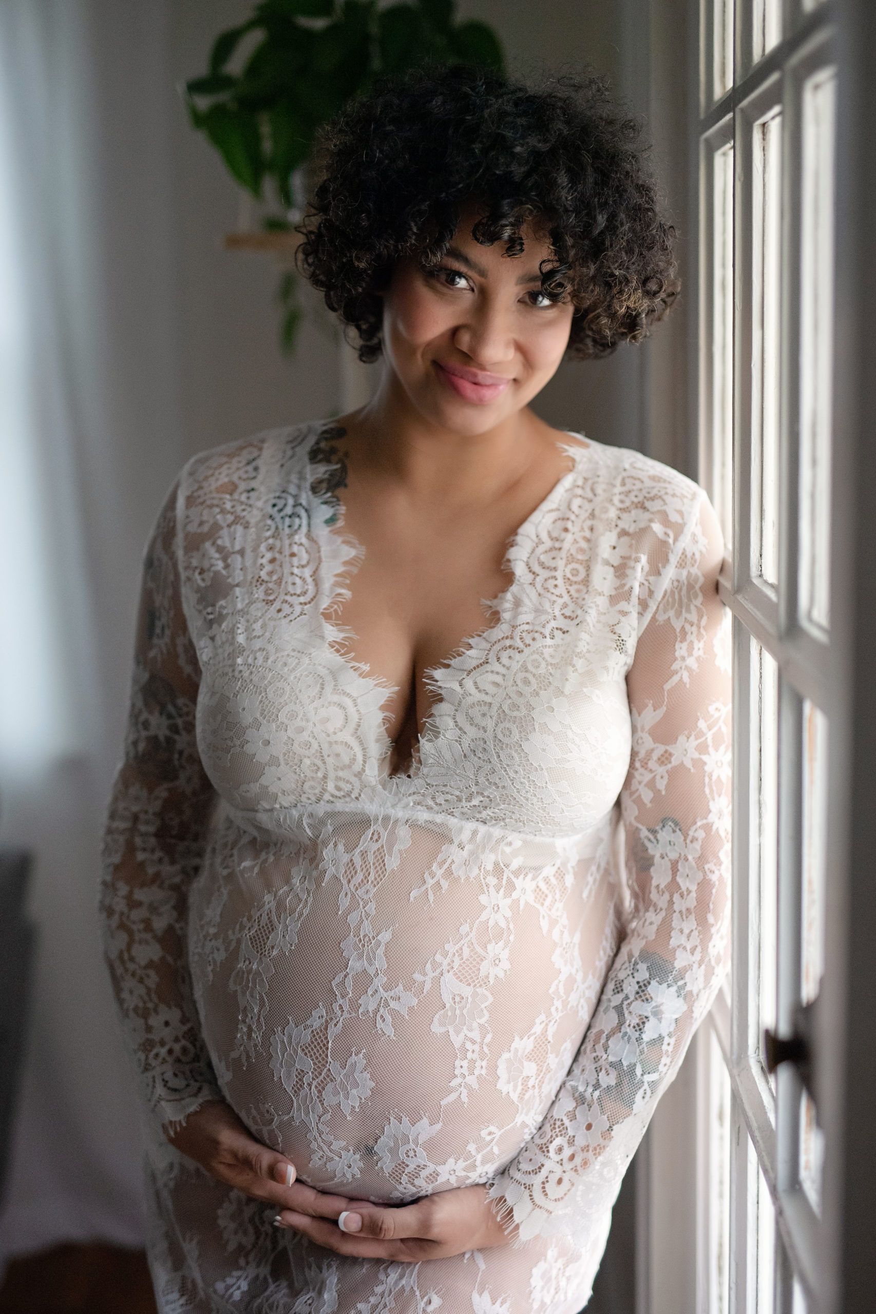 Pregnant woman in white lace dress, smiling, standing near window.
