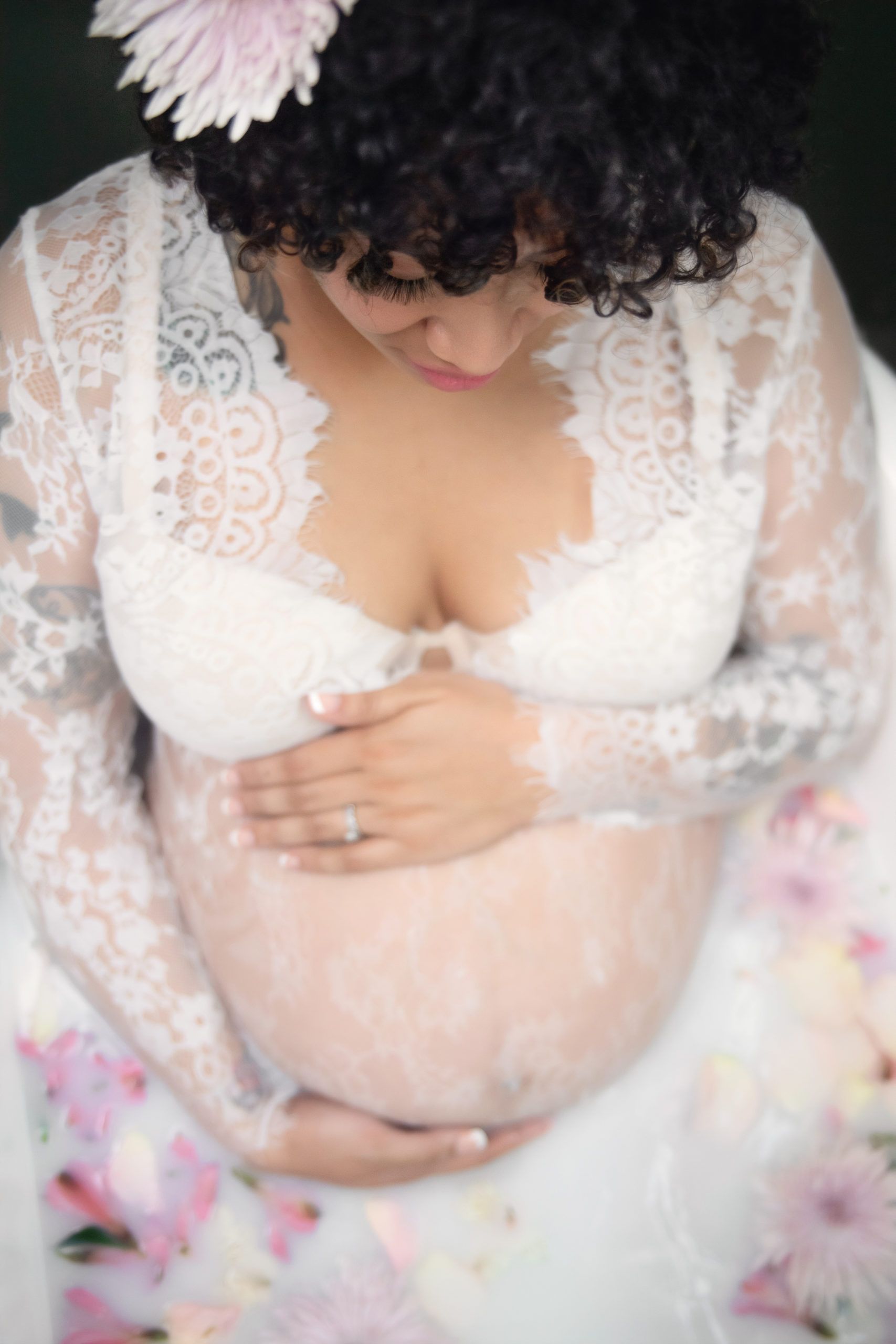 Pregnant woman in lace gown cradling belly, looking down, flower in hair.