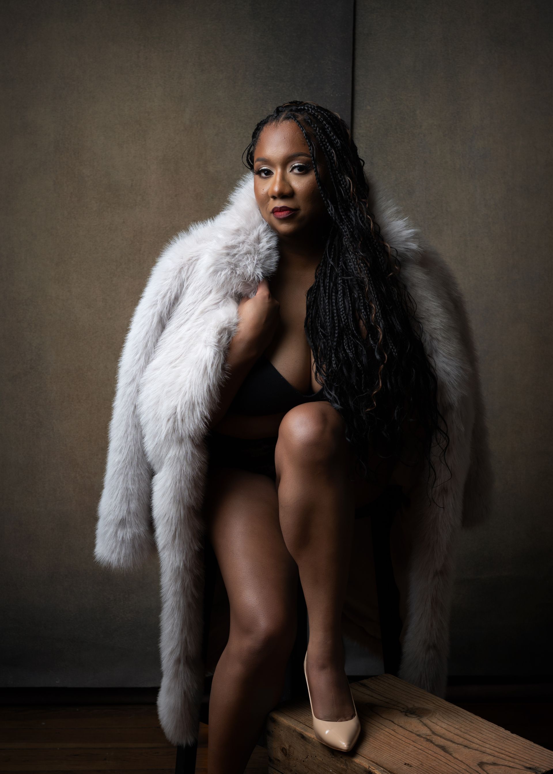 Woman in fur coat, black lingerie, sitting on wood, beige heels, long dark braids.