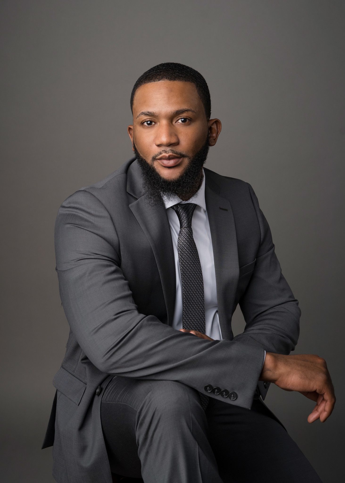 Man in gray suit, seated, looking at the camera, neutral expression. Gray background.