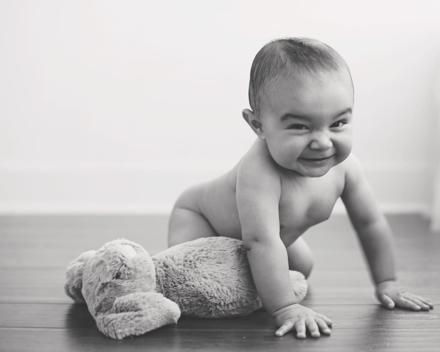Smiling baby crawling on a wood floor, leaning over a plush toy. Black and white.