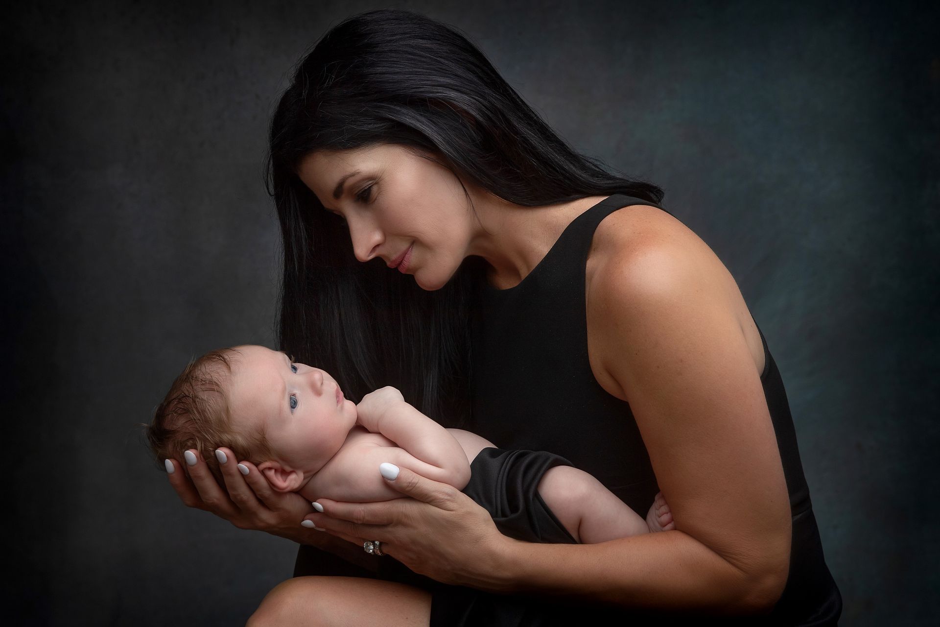 Mother with dark hair gazing at her baby, cradled in her arms, against a dark background.
