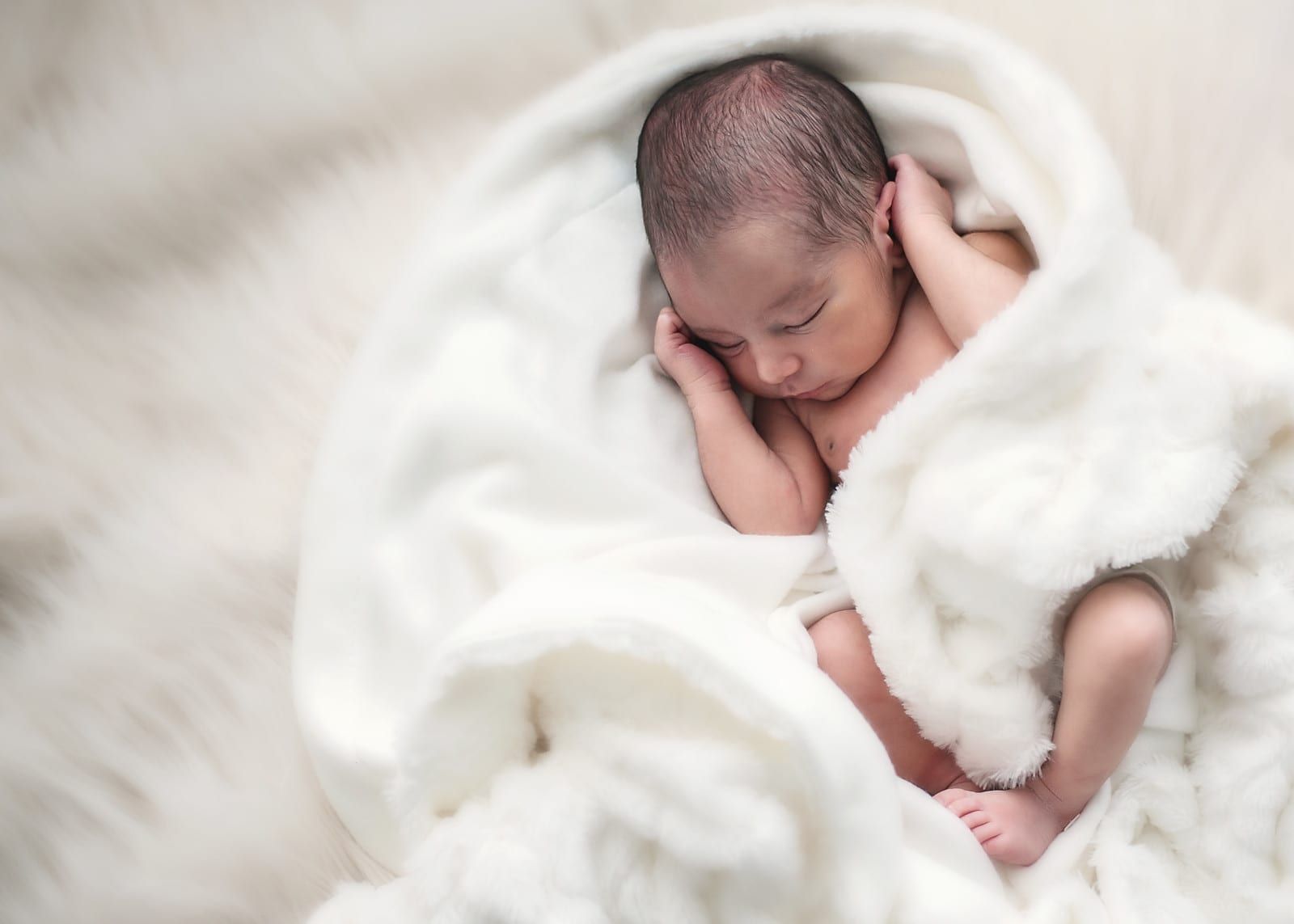Newborn baby wrapped in a fluffy white blanket, sleeping peacefully with hands near face.