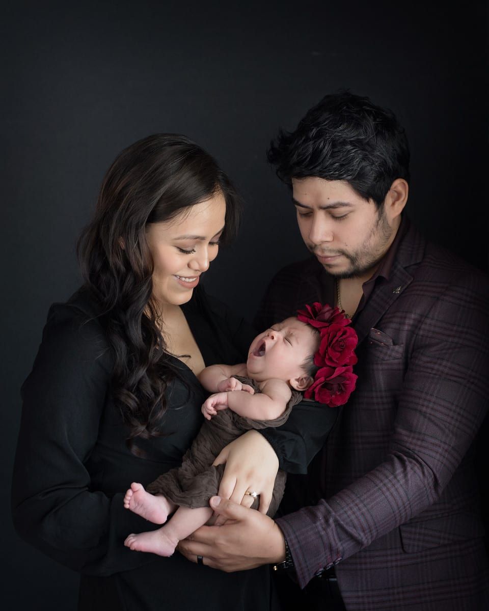 Parents holding a newborn baby wearing a floral headpiece, smiling against a black background.