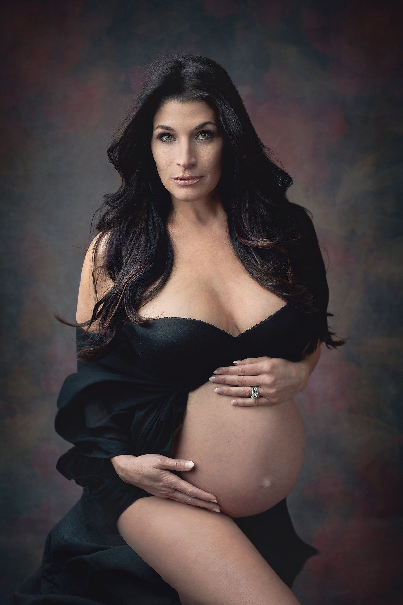 Pregnant woman in black dress, holding her belly, with dark hair and backdrop.