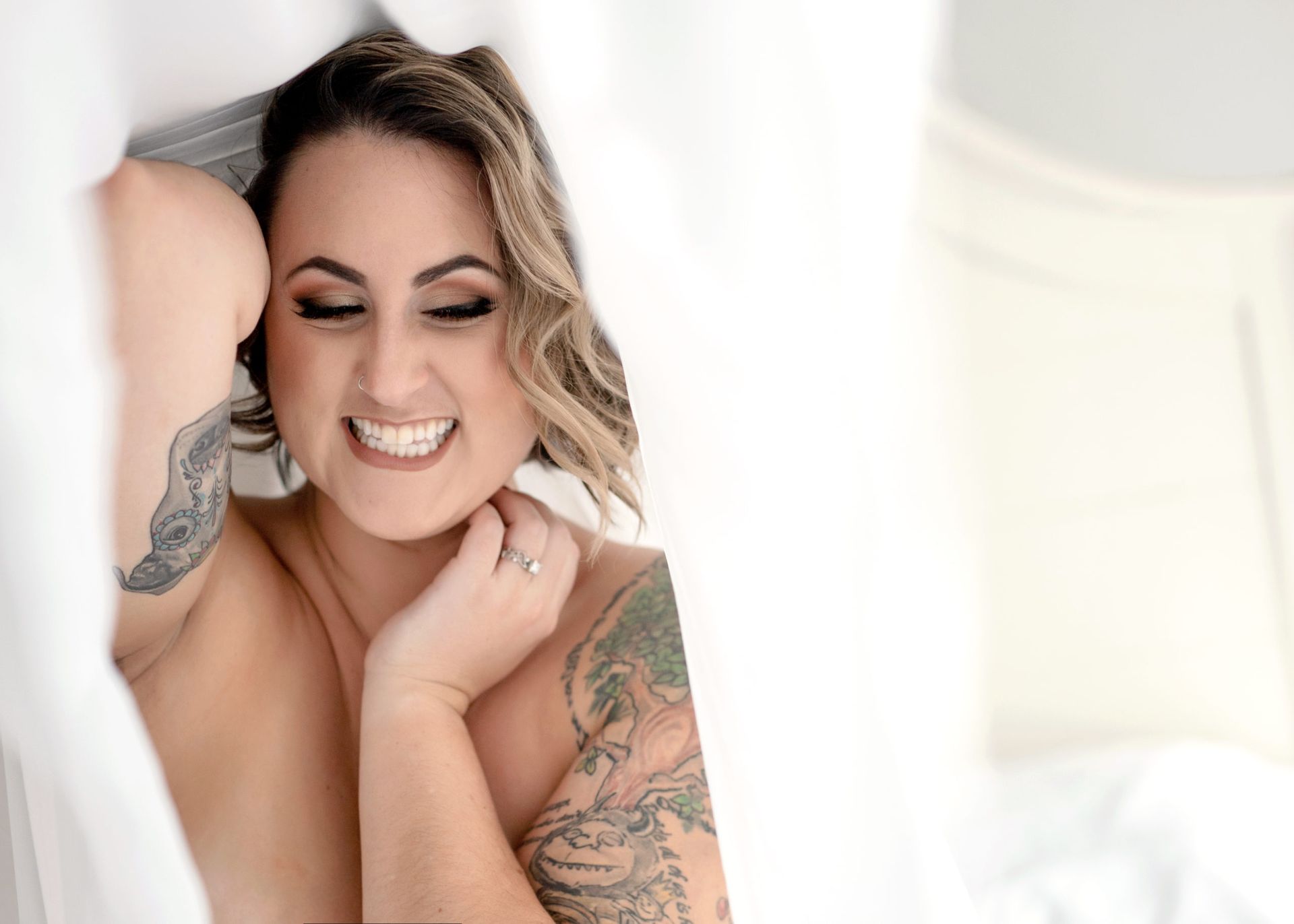 Woman with tattoos, smiling, peeking out from behind sheer white curtains.