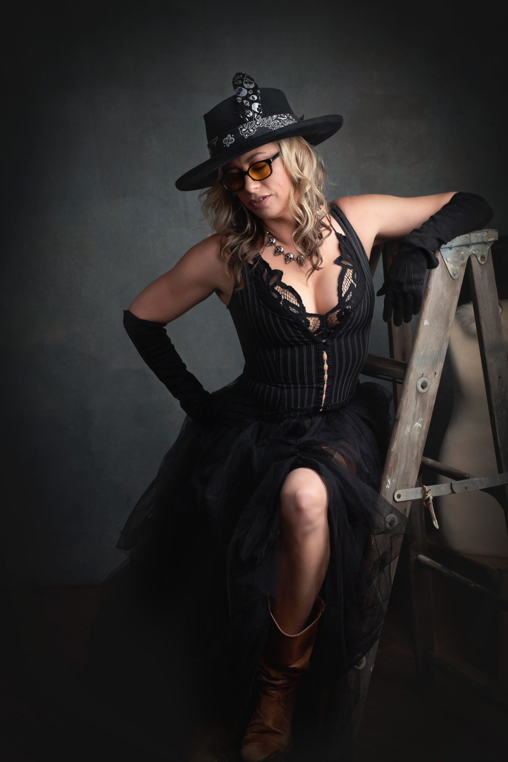 Woman in black outfit and cowboy boots, leaning on a ladder.