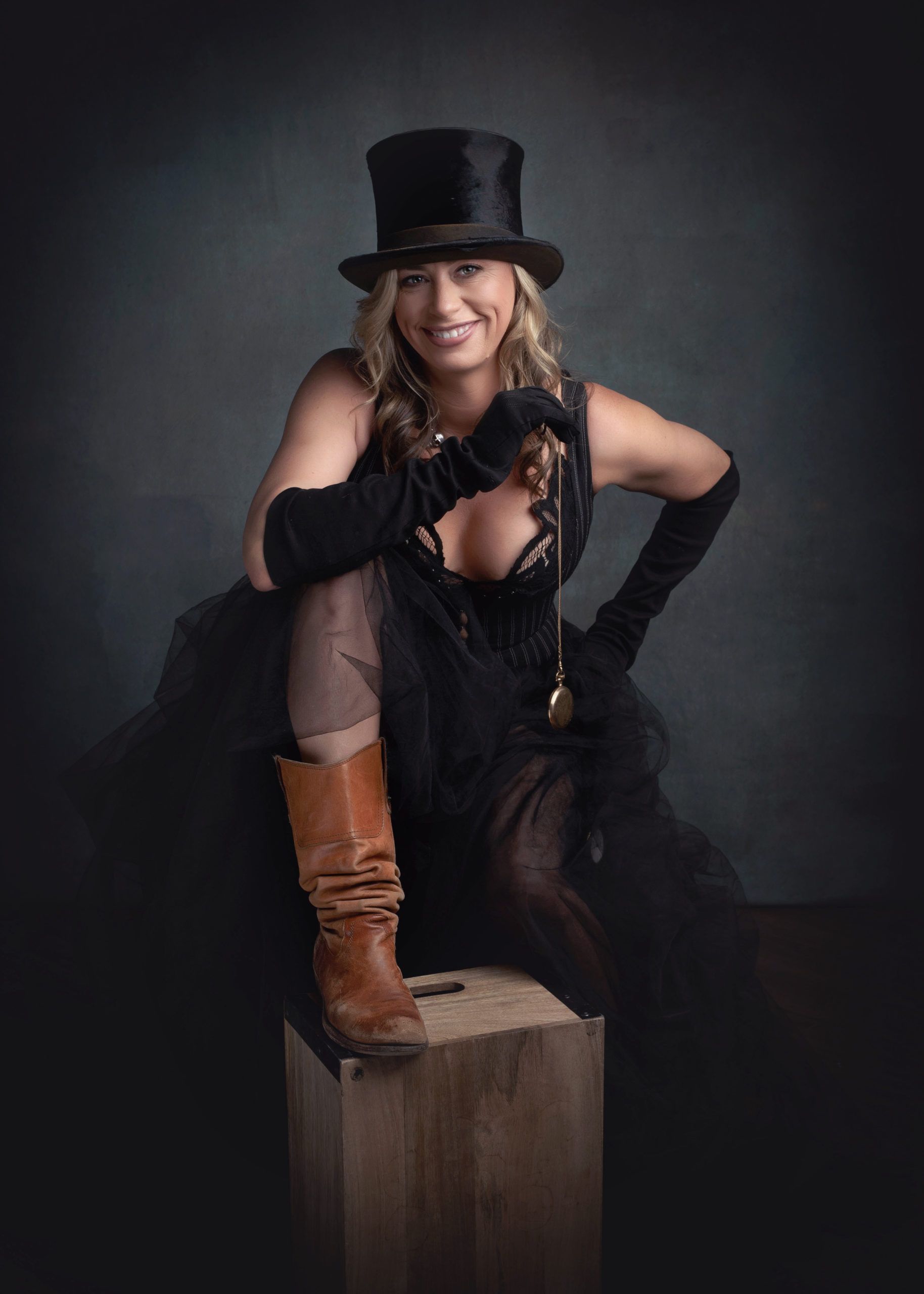 Woman in steampunk attire, wearing a top hat, boots, and gloves, poses confidently on a wooden box against a dark background.