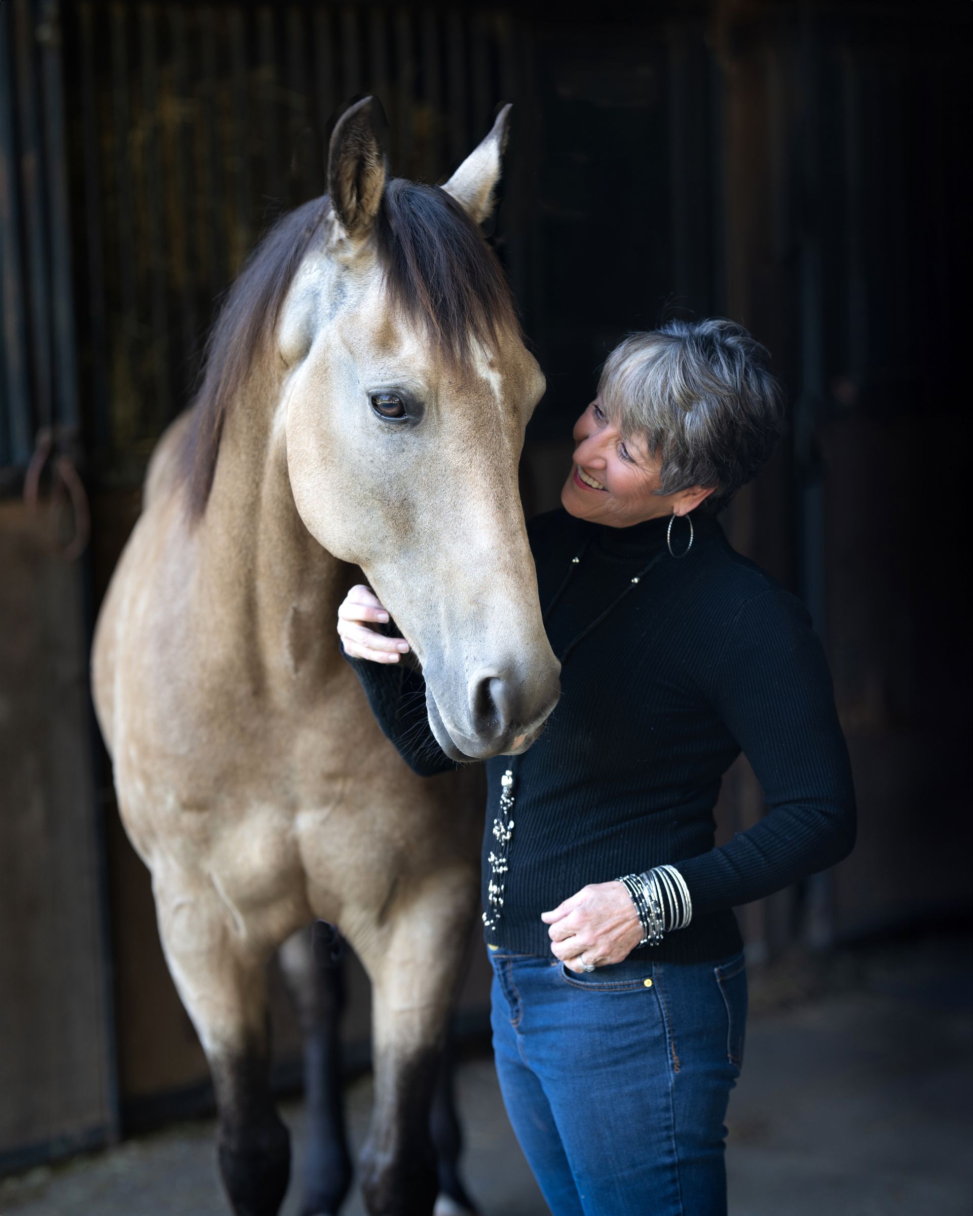 Woman in black sweater smiles at a tan horse in a stable.