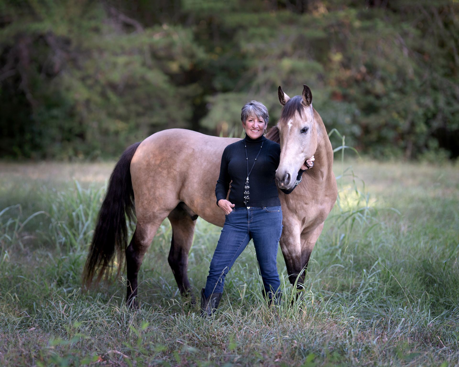 Woman in jeans and black top, posing with a tan horse in a grassy field.