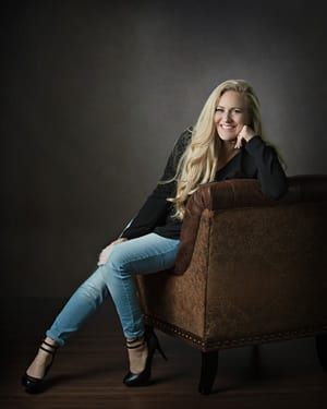 Woman with long blonde hair, smiling, sits in armchair, wearing jeans and black top, heels.