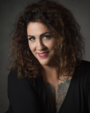 Woman with curly brown hair, smiling, wearing a black top, and has a tattoo on her upper chest.