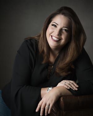 Woman with brown hair smiles, leaning on armrest. She wears a black blouse, looking at the camera.