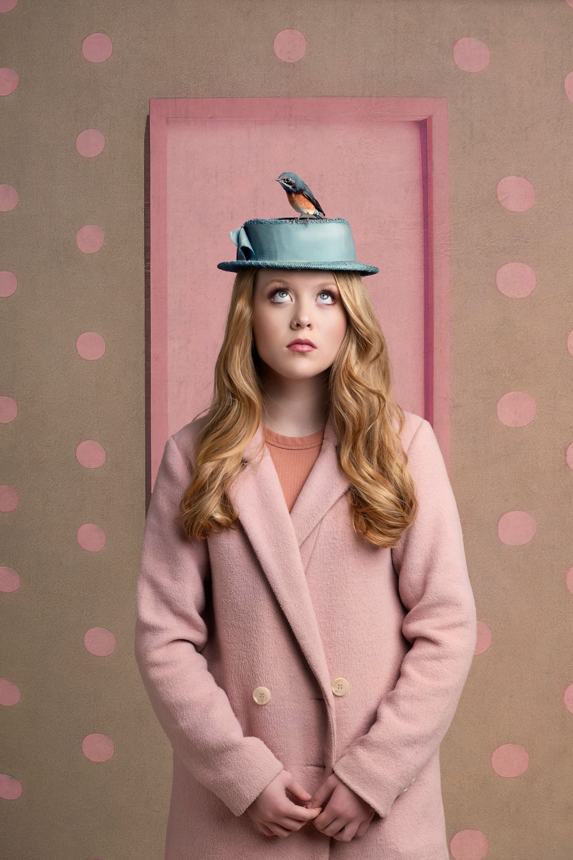 Blonde woman in pink coat and hat with a bluebird perched on top, looking up, pink door backdrop.