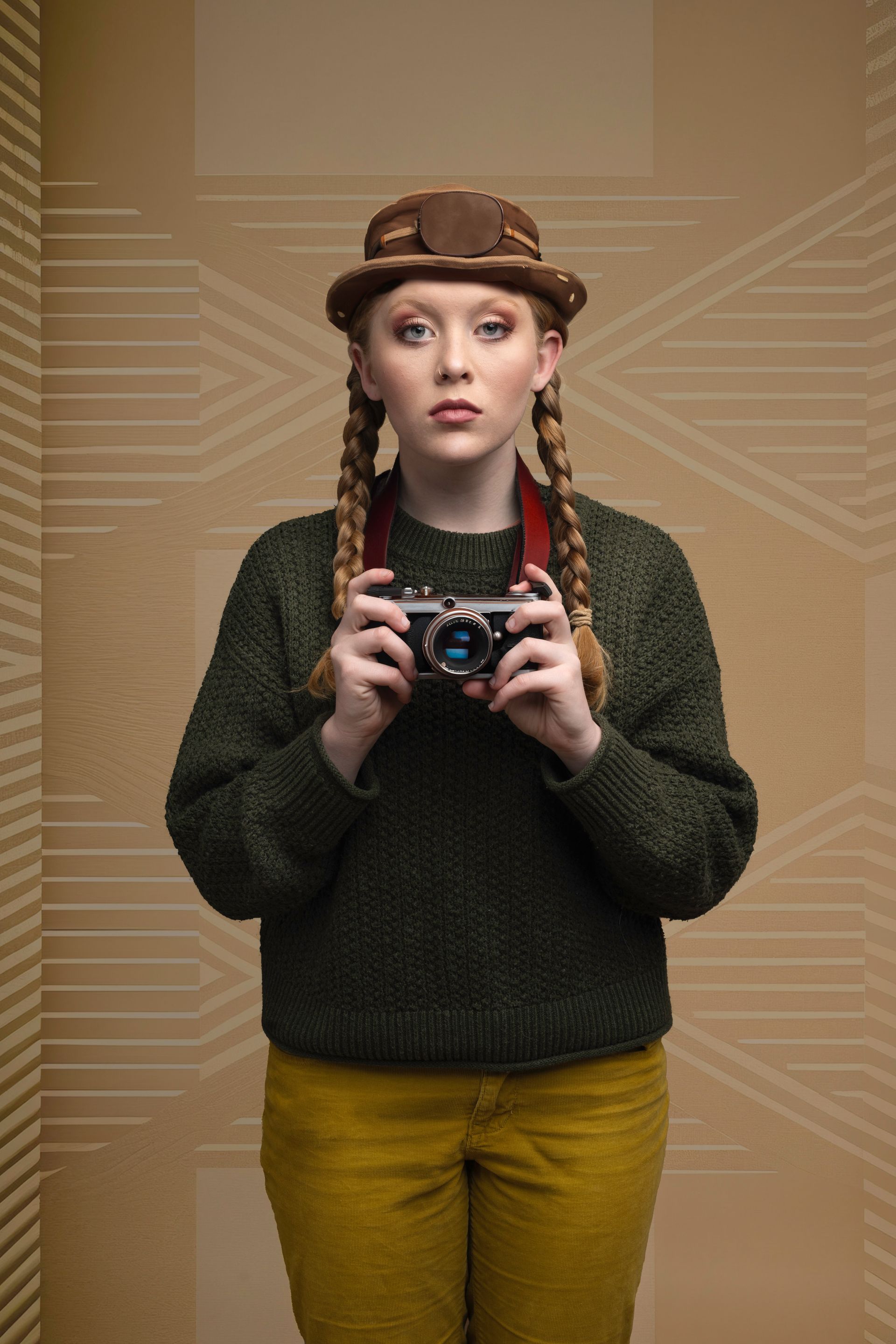 Woman in green sweater, yellow pants, and hat holds a camera. Braided hair, neutral background.