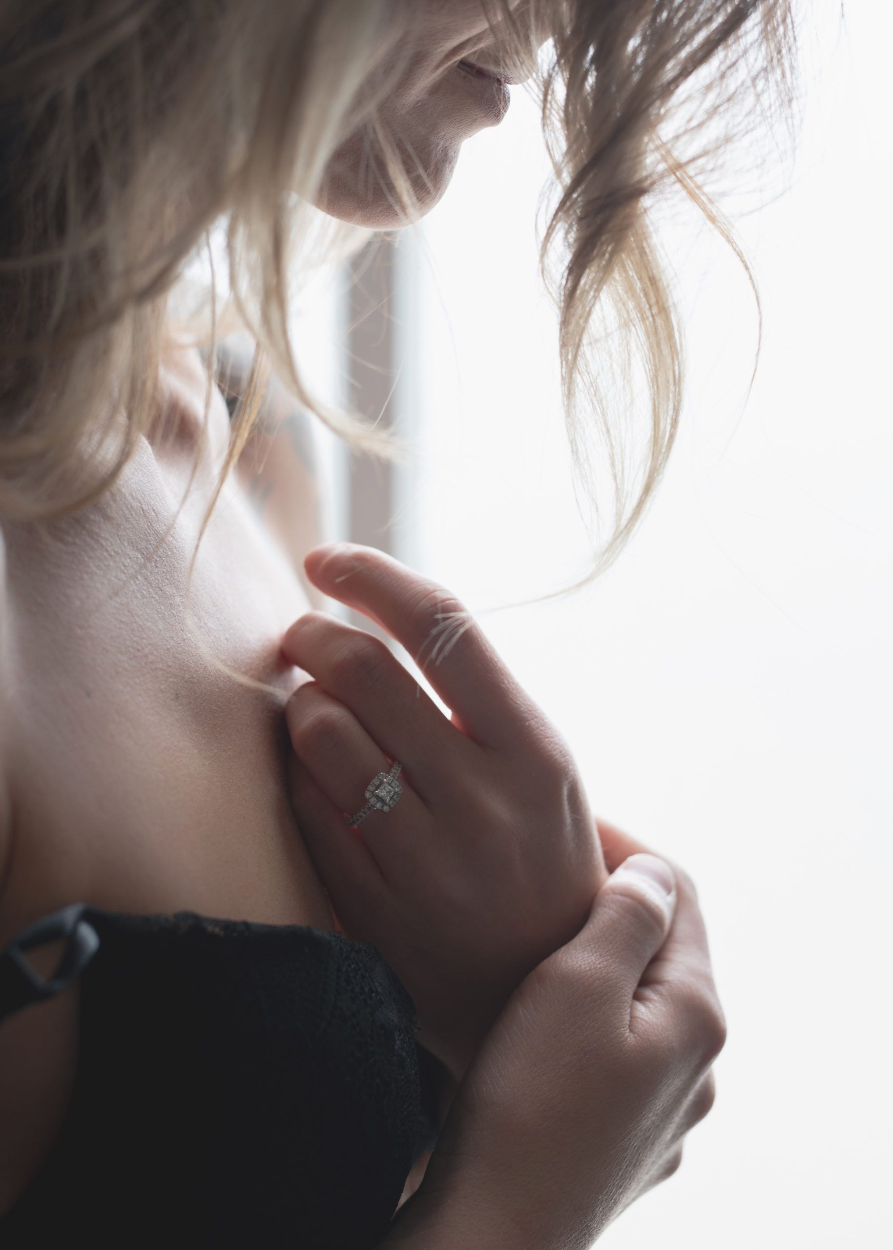 Woman in black lingerie, hands on chest, wearing a ring. Blonde hair, soft light.