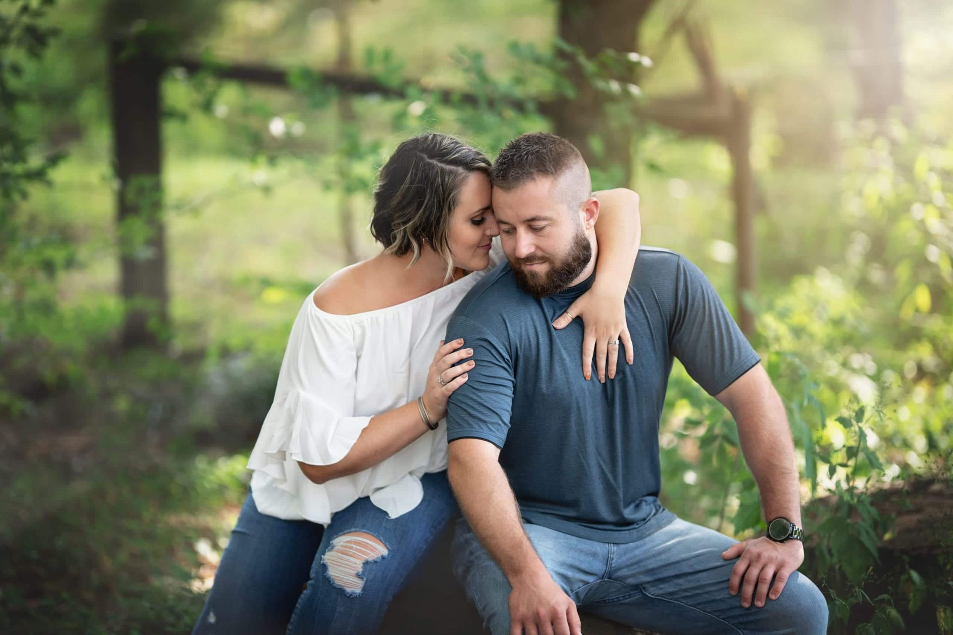 Woman kisses man's head, both seated outdoors. She wears white top, he has beard and blue shirt.